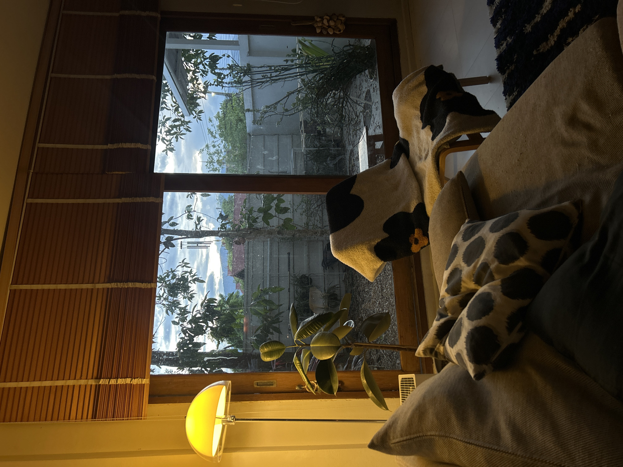 An interior scene is captured, possibly in Chang Phueak, Thailand, as hinted in the prompt. A window dominates the view, showcasing the outdoors with trees and possibly residential structures beyond. The window has wooden blinds on the left. A floor lamp stands in front of the window on the lower part of the frame. To the right of the window, there's a chair draped with a blanket featuring a pattern, along with a portion of a sofa or a chair covered with pillows. The overall lighting suggests it may be evening or a dimly lit space.