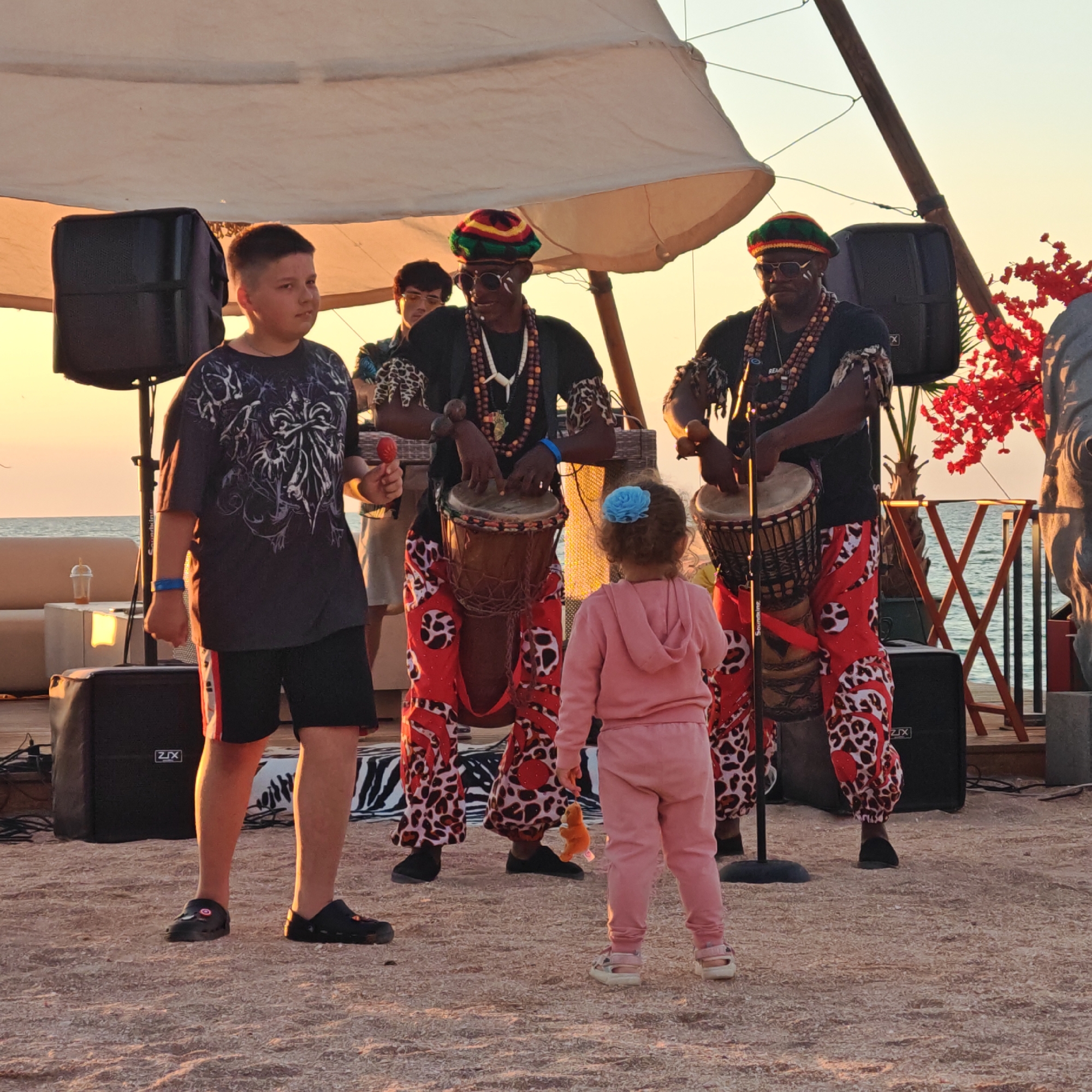 This image depicts an outdoor performance with a group of musicians, likely a drum ensemble, on a sandy ground near a body of water, possibly the Sea of Azov. In the foreground, a young boy stands facing the performers, holding a red maraca and wearing a dark t-shirt and black shorts. To his right, a young girl in a pink tracksuit faces away from the camera, also holding a small orange toy. The performers, dressed in vibrant, patterned trousers and traditional headwear, are actively playing large drums. One of the musicians, in the center, is also wearing beads around his neck and has white face paint. In the background, a large white canopy is stretched out, providing shade. Two black speakers are visible, one to the left and another to the right of the performers. A red artificial plant with bright red flowers is positioned to the right of the stage. The sky is a soft orange, suggesting it is either sunrise or sunset. The environment suggests a coastal location, possibly a beach or boardwalk setting within the city of Shchelkino, Ukraine.
