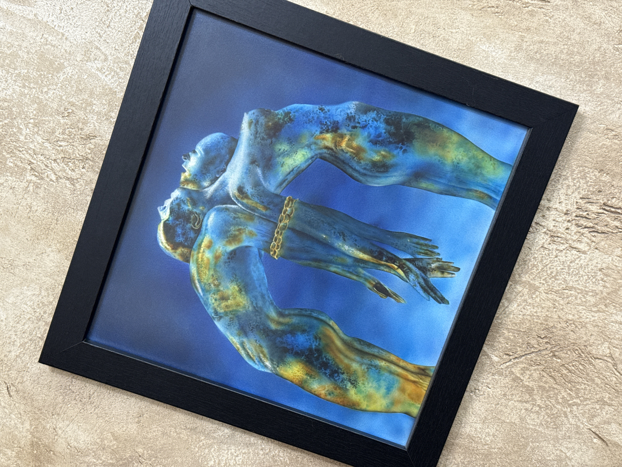 A framed image of two blue-toned human figures is positioned on a textured surface with a light brown hue. The frame is black, and the image depicts the figures in a symmetrical, possibly symbolic, arrangement. The location, Burgas, Bulgaria, is not evident in the image.
