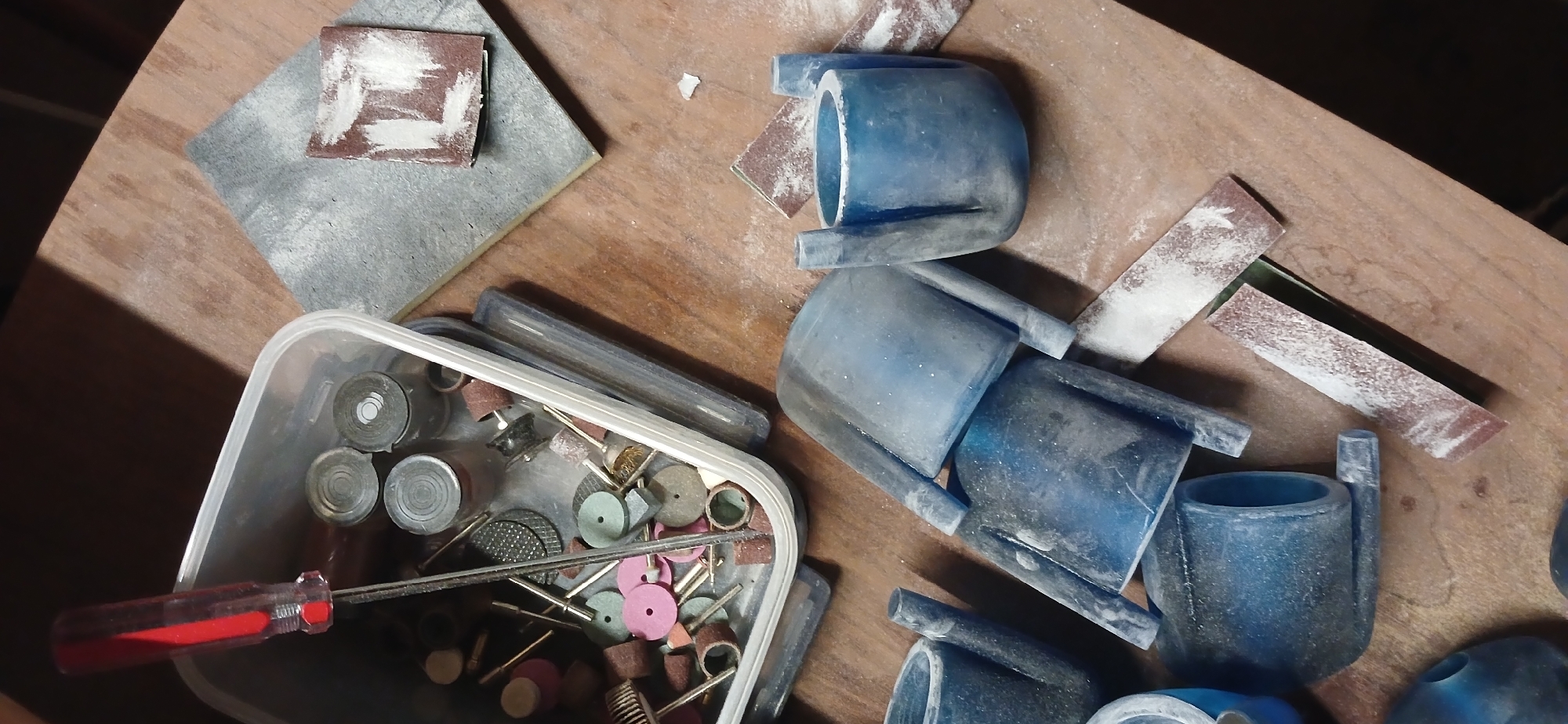 This image presents a close-up, overhead view of a dusty workbench, characteristic of a crafting or workshop environment. The surface is light brown wood, heavily speckled with fine white dust, indicating recent or ongoing work.
Numerous tools and materials are visible. Scattered across the wooden surface are several pieces of used sandpaper, also coated in white dust, alongside a larger, rectangular grey abrasive pad in the upper left.
The prominent subjects on the right side of the frame are multiple blue-colored, molded objects. These items, likely made of plastic or resin, resemble curved pipe sections or handles with tubular extensions. They are also covered in the same white dust, suggesting they are undergoing a finishing process like sanding or grinding. Some are stacked, while others lie individually.
In the lower-left, a clear plastic container holds a diverse collection of small rotary tool accessories. This includes various sanding drums, abrasive grinding bits, small circular polishing wheels in shades of pink, grey, and brown, and miniature wire brushes, all designed for detailed work. A red-handled screwdriver or similar small tool is partially visible within the container.
The overall scene depicts an active crafting space dedicated to shaping and refining materials, likely using rotary tools and abrasive methods. No people are present, and there is no visible text. The image is a detailed snapshot of a workspace and offers no specific visual cues to its reported location in Moscow, Russia.