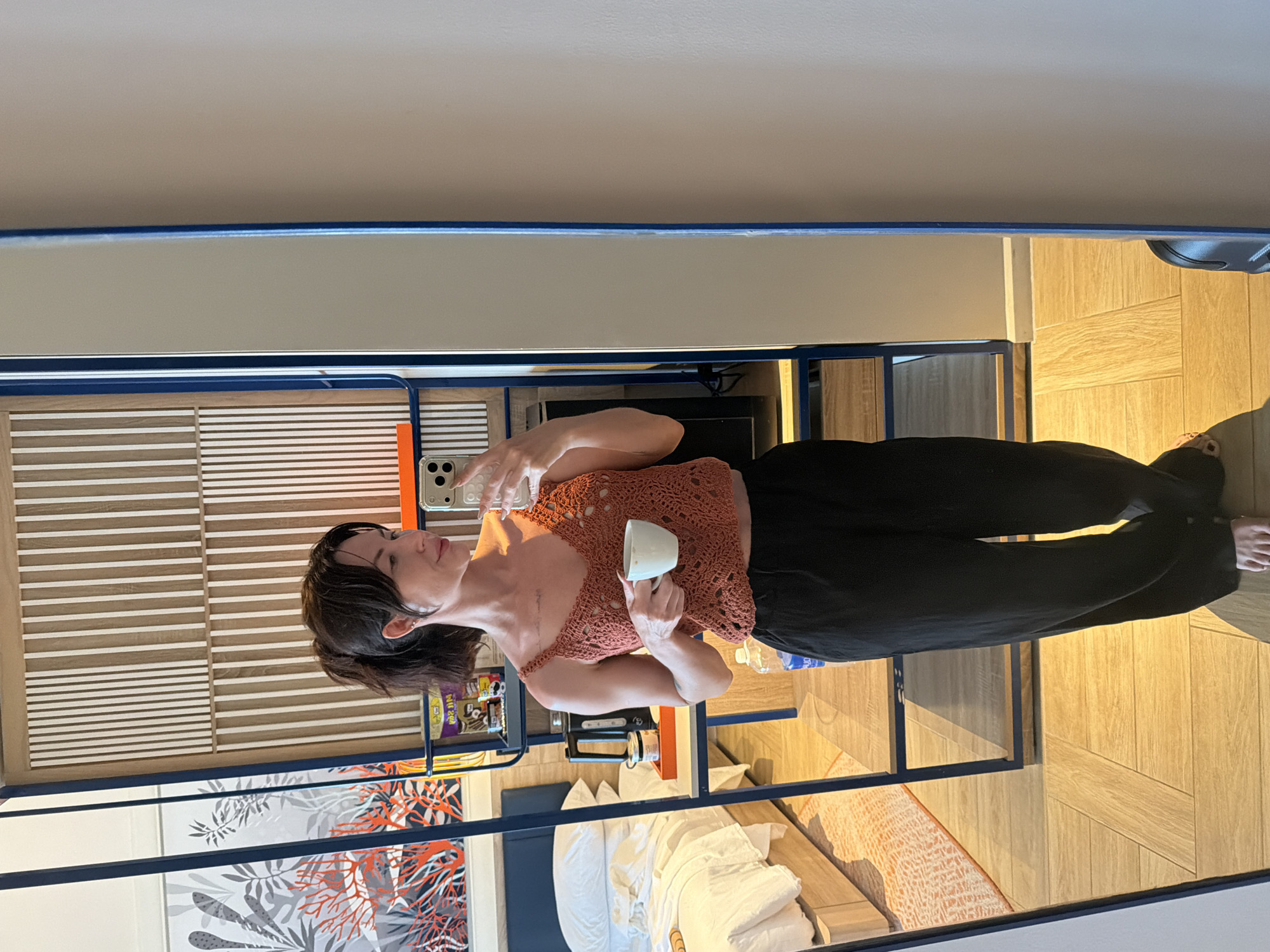A woman is standing in a hotel room, holding a cup of coffee and taking a mirror selfie. She is wearing a crochet top and black pants. The room has a bed, a mirror, and a shelf with various items. The floor is made of wood. The image is taken from a medium distance, showing the woman from the waist up, with the background reflected in the mirror. The overall lighting is bright and natural.