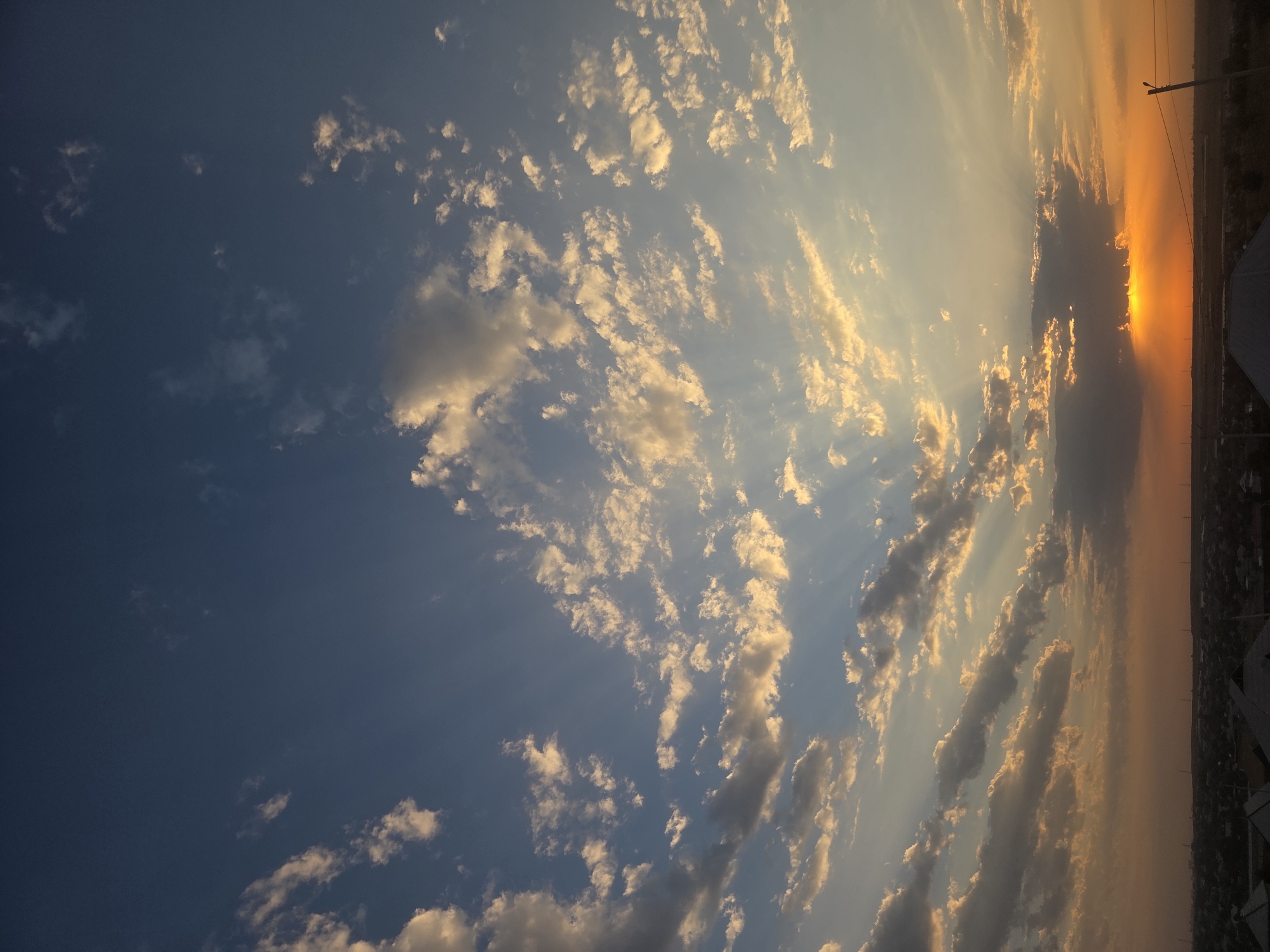 The image shows a wide shot of the sky above a city at dusk. The sky is a gradient of deep blue at the top, transitioning to lighter blue and then orange near the horizon. Scattered clouds are illuminated by the setting sun, creating dramatic shafts of light. The sun itself is a bright orange sphere just above the horizon, partially obscured by dark clouds. Below the sky, a dark silhouette of a city is visible, with rooftops and some indistinct structures. The overall impression is a serene and picturesque sunset over an urban landscape in Қарасу, Kazakhstan. There are no people or specific actions depicted in the image.
