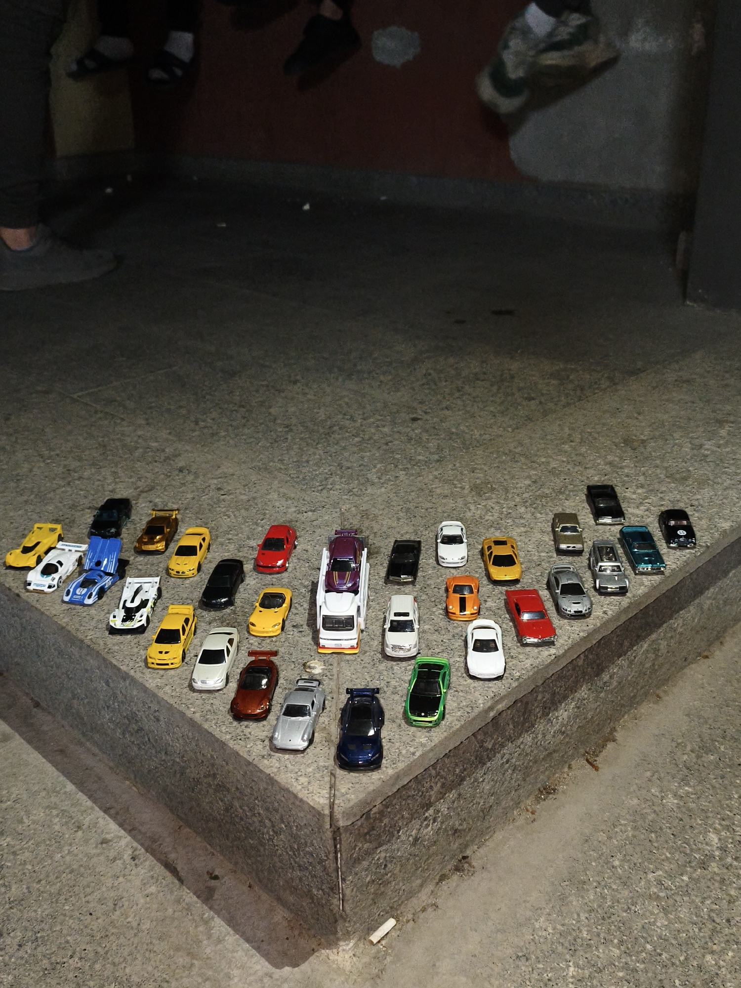 The image shows a collection of toy cars arranged on a low concrete ledge, possibly a step or a curb, in an urban environment. The collection features a variety of colors and models, including sports cars, sedans, and a car carrier. The ledge is made of grey concrete, and the ground around it is a similar grey material. The lighting is subdued, casting shadows across the scene. Several people are partially visible in the background, with only their legs and feet showing. They are wearing various types of footwear, including sneakers and sandals. The background suggests an indoor or sheltered area, possibly a doorway or entrance to a building, with a dark wall in the upper part of the image. There are also a few small pieces of litter on the ground near the ledge. The image does not provide information to determine the city of Unknown.