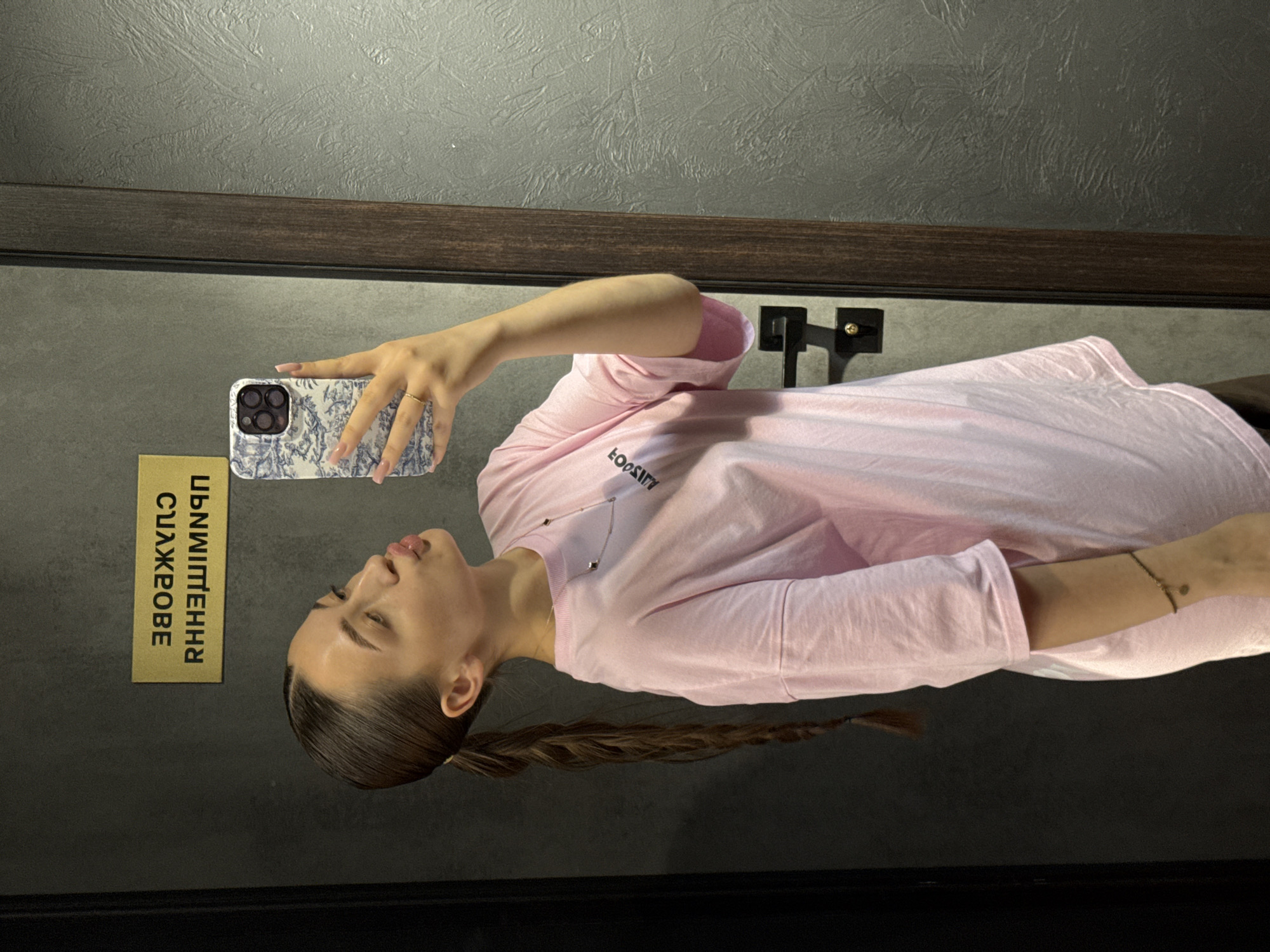 A young woman is taking a mirror selfie in a room with grey textured walls and a wooden shelf. She is wearing a pink long-sleeved t-shirt with a small silver necklace. Her hair is dark brown and pulled back into a braid. She is wearing a ring on her left hand and a bracelet on her right wrist. She is holding a white phone case with a blue and white pattern. To her left, there is a gold sign with text in Ukrainian, which reads "СЛУЖБА ЗДОРОВ'Я". The room appears to be an indoor space, possibly a dressing room or a bathroom. The lighting suggests it could be daytime or artificial light.