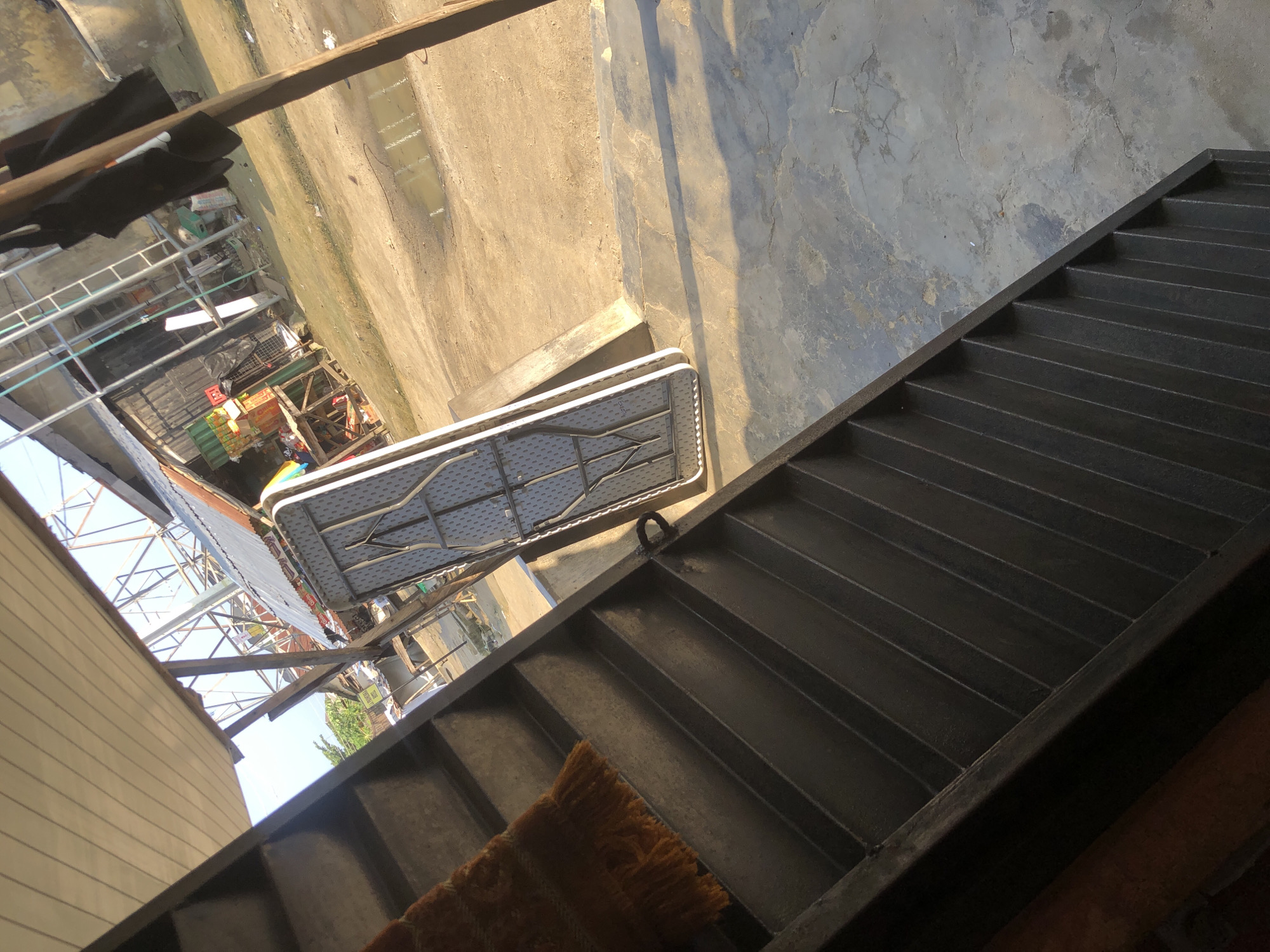 The image depicts an outdoor scene that appears to be a market area in Ado, Nigeria. In the foreground, there is a dark, ribbed staircase, partially covered by a brown and orange striped rug with fringe. To the right of the staircase, a folded white plastic table with metal legs is propped against a concrete structure. The background reveals a dusty, uneven ground, with a small puddle of water visible. Beyond the ground, there are various structures including corrugated metal roofs, wooden stalls, and what looks like a metal scaffolding. Several items are visible on the stalls, including crates of goods, bags, and possibly some fabric. The overall impression is one of a bustling, possibly informal, marketplace environment under a bright sky.