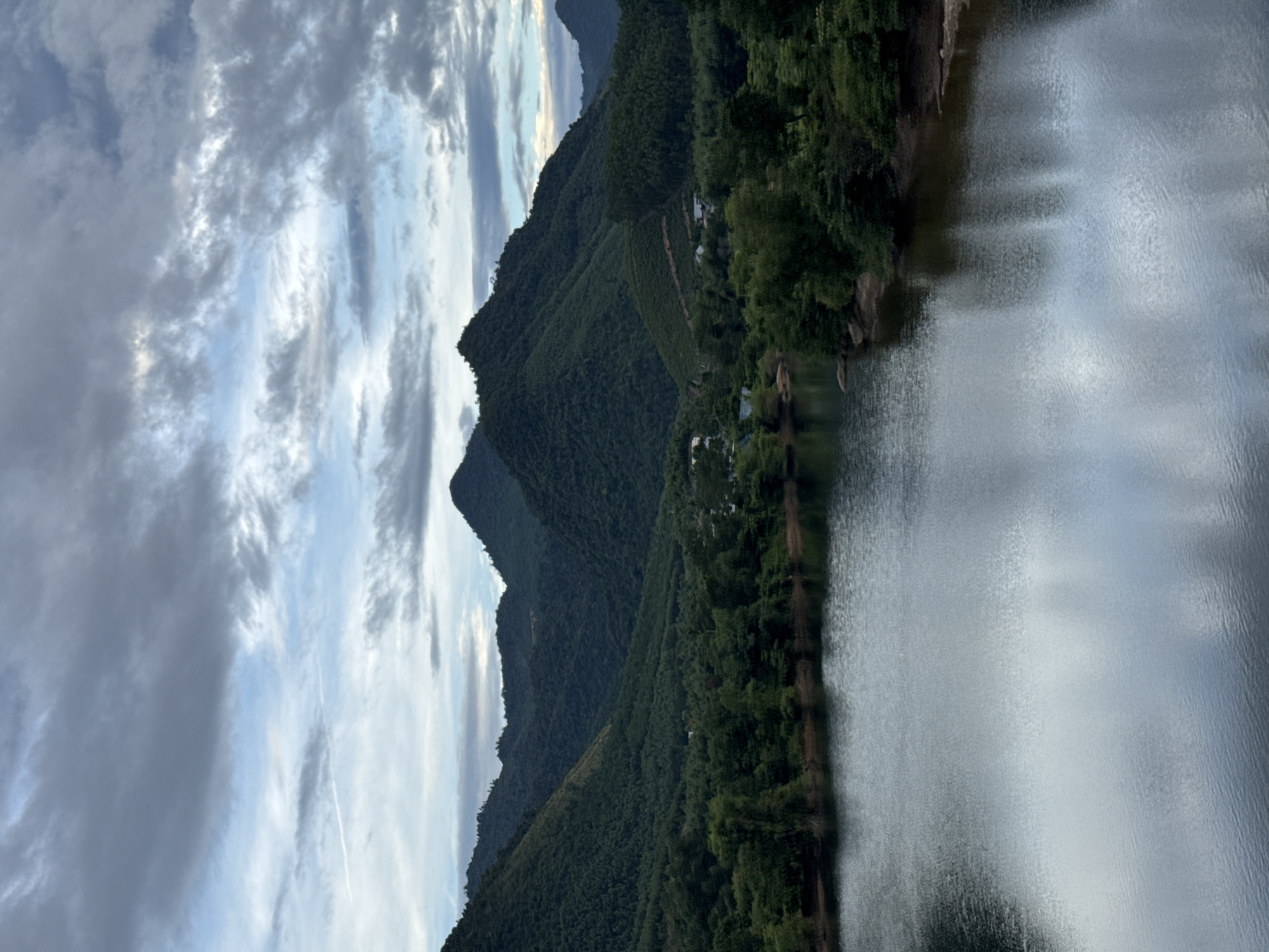 The image depicts a tranquil scene of a lake in the foreground, reflecting the overcast sky and surrounding lush green mountains. The mountains are densely covered in vegetation, with varying shades of green indicating different types of trees and foliage. Some scattered houses and buildings are visible nestled amongst the trees on the slopes of the mountains, suggesting a rural or semi-rural setting. The water of the lake is dark and still, with subtle ripples disturbing its surface. The sky is filled with dramatic clouds, with patches of lighter blue peeking through, hinting at a possibly overcast or twilight condition. There are no people or animals visible in the frame. The overall atmosphere is serene and natural.
While the image showcases a beautiful natural landscape, it is not possible to definitively identify its specific location within the city of Đà Nẵng, Vietnam, based solely on the visual information provided. Đà Nẵng is known for its beaches and urban development, but also has surrounding mountainous areas and reservoirs that could potentially resemble this scene.