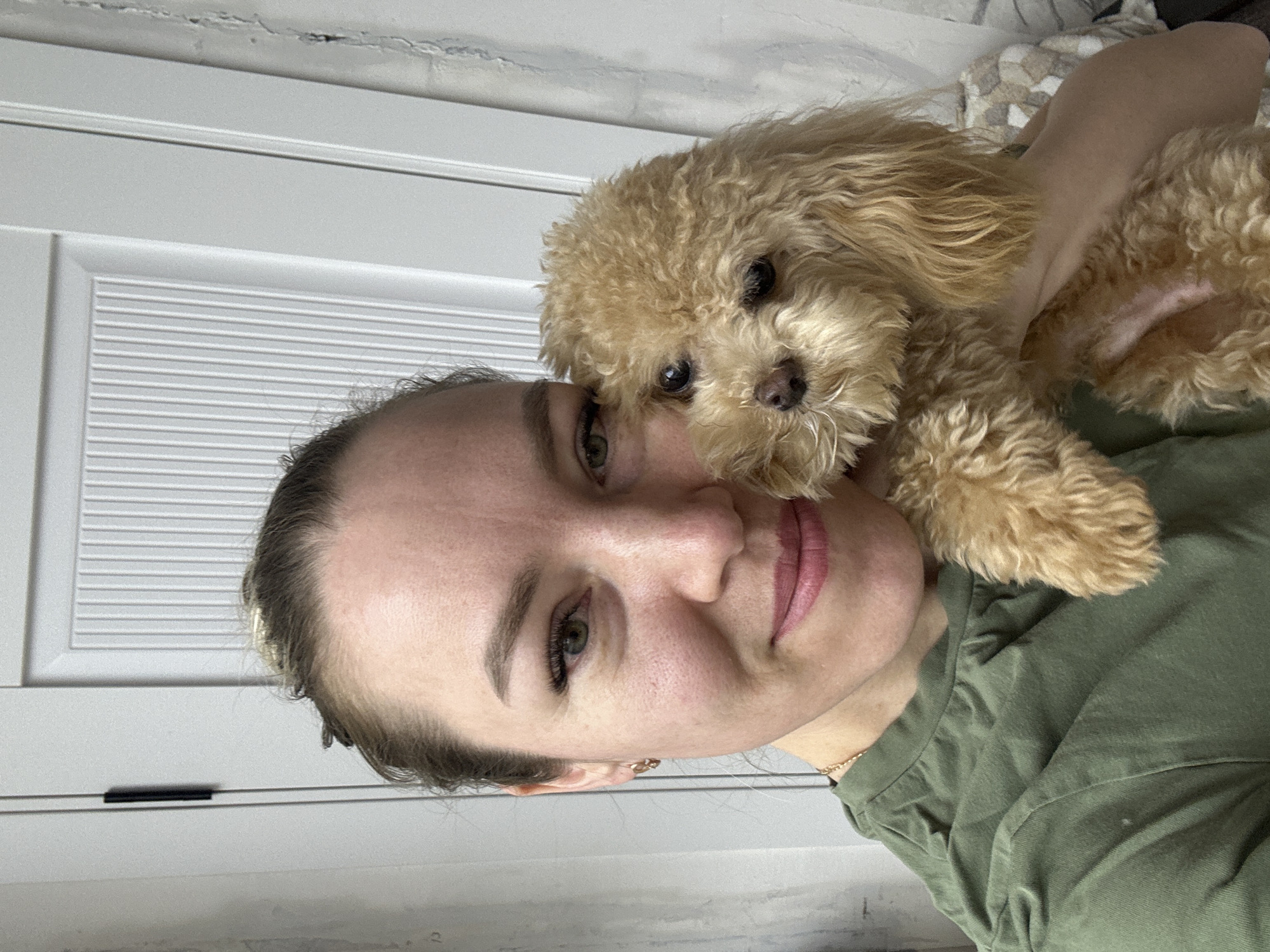 A fair-skinned woman with her hair pulled back is holding a small, light-brown poodle in her arms. The woman is wearing a green shirt, and a gold chain is visible around her neck. She has light eyebrows, eyelashes, and pink lips. Her green eyes are looking directly at the camera. The dog has curly fur and is looking at the camera as well. The background consists of a white door with horizontal panels and what appears to be a patterned blanket or bedsheet. The location cannot be accurately confirmed as being in слобода Старая, Russia without additional context.