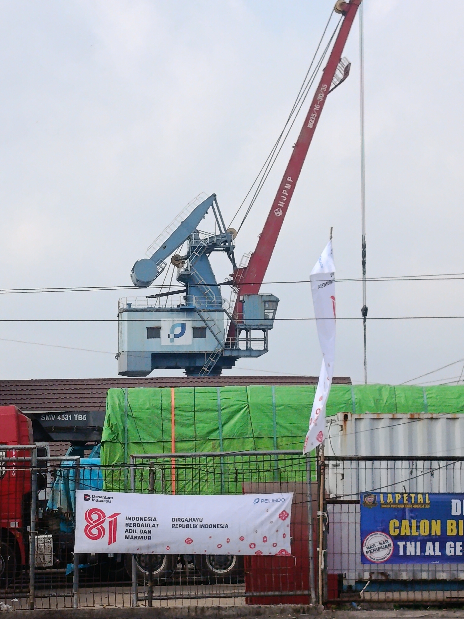 The image captures a port scene with industrial cranes and cargo containers. A large red crane with markings "NJ PMP" and "MQ35/16-30/35" is prominent, alongside a blue crane with a "J" logo. In the foreground, a banner reads "81 INDONESIA BERDAULAT ADIL DAN MAKMUR DIRGAHAYU REPUBLIK INDONESIA," and another banner displays "LAPETAL CALON BI TNI. AL GE." Trucks, one partially visible and red, are also present, along with a cargo container covered in green tarpaulin. The overall environment suggests a busy logistics or shipping hub. The presence of the banner with "DIRGAHAYU REPUBLIK INDONESIA" and the specific reference to "TNI. AL" (Tentara Nasional Indonesia - Angkatan Laut, Indonesian National Army - Navy) indicates a connection to Indonesian national identity and potentially a military port. While the image depicts a port, the specific location in Palembang, Indonesia cannot be definitively determined from the visual information alone.