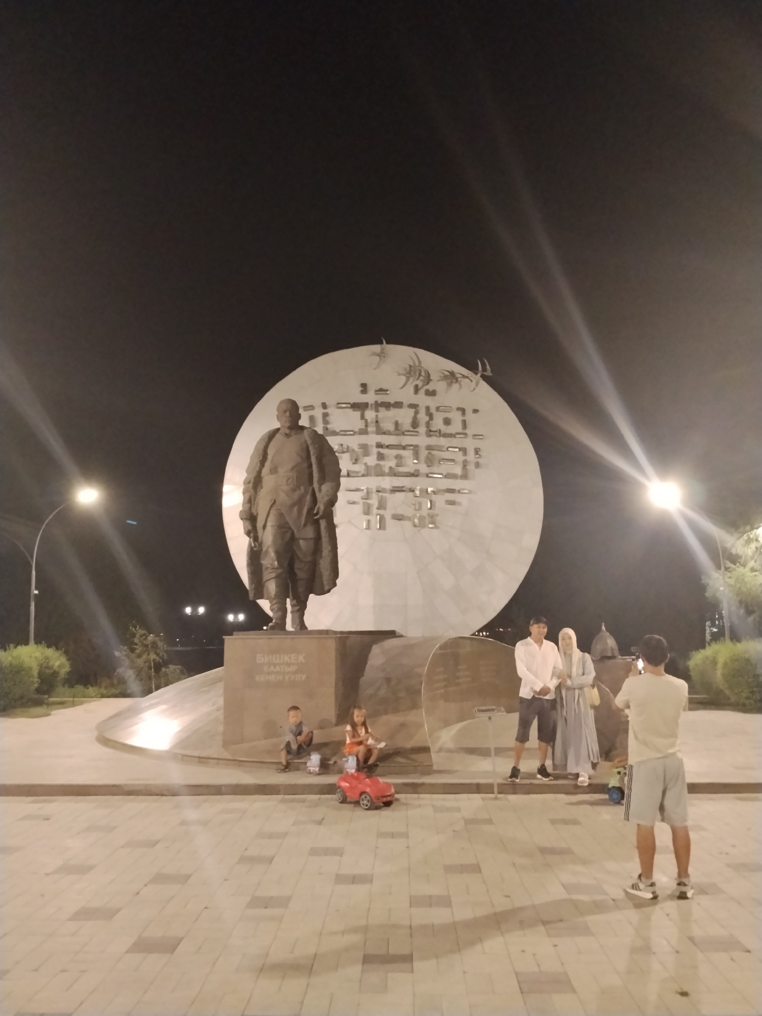 The image captures a nighttime scene at a monument in Bishkek, Kyrgyzstan. In the foreground, a man stands facing away from the camera, holding a phone and appearing to take a photograph. To his left, a couple stands posed, with the man wearing a white shirt and dark shorts, and the woman in a light blue headscarf and dress. Behind them, a large circular monument features intricate metallic designs. To the left of the monument, a bronze statue of a man in a fur-lined coat stands on a pedestal. In front of the pedestal, two young children are seated, with a red toy car nearby. The surrounding area is paved with patterned tiles, and trees and streetlights are visible in the background. The sky is dark, with light beams from streetlights creating a dramatic effect. The text on the pedestal appears to be in Cyrillic script and reads "БИШКЕК БААТЫР КЕНЕН УУЛУ", which translates to "Bishkek Baatyry Kenen uulu".