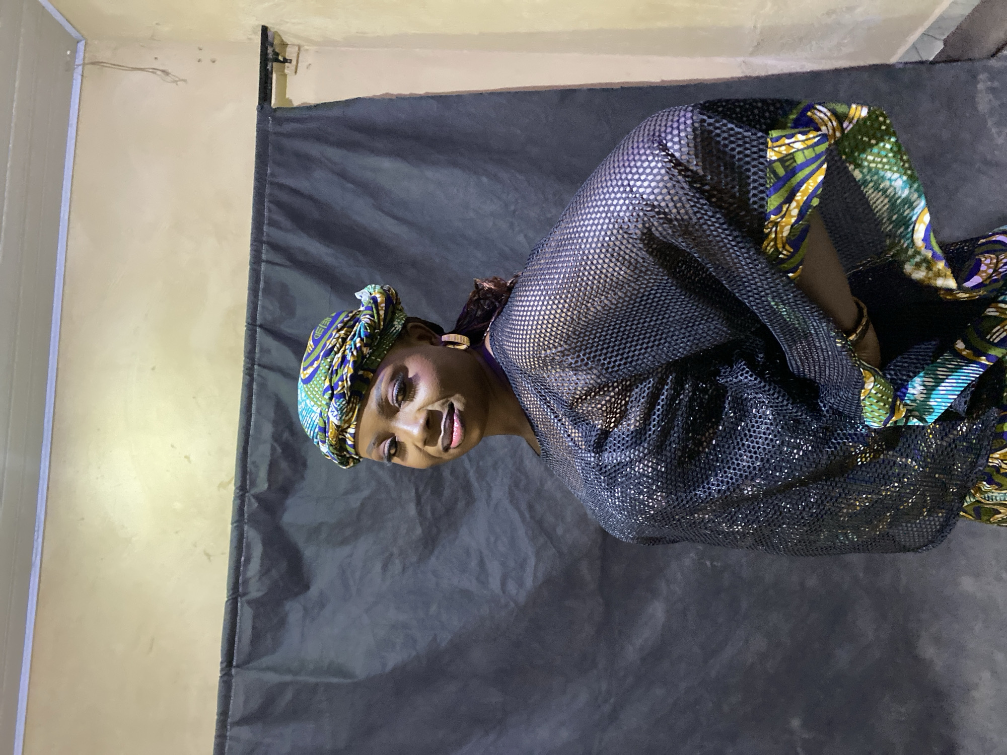 A portrait photograph captures a woman, likely of African descent, posing directly for the camera with a subtle, pleasant smile. She appears to be in her 20s or 30s.
The woman is adorned in a vibrant, multicolored headwrap featuring patterns in green, blue, yellow, and white. Her top is a dark, seemingly metallic mesh or sparkly fabric, beneath which a patterned material, possibly matching her headwrap, is visible. She wears simple gold-toned hoop earrings and a thin gold-toned bracelet on her right wrist.
The setting appears to be an indoor, possibly makeshift studio. The main background behind her is a dark grey or black fabric backdrop, showing some wrinkles. To the left of the backdrop, a light tan or off-white wall is visible, complete with some minor surface imperfections and what looks like thin wiring near its upper edge. A dark vertical element, potentially a door frame or a structural support, can be seen on the far left.
The lighting is focused on the subject, illuminating her face and attire. The overall mood conveyed is calm and confident. There is no discernible text visible in the image. The location context provided is Kuru Dungus, Nigeria.