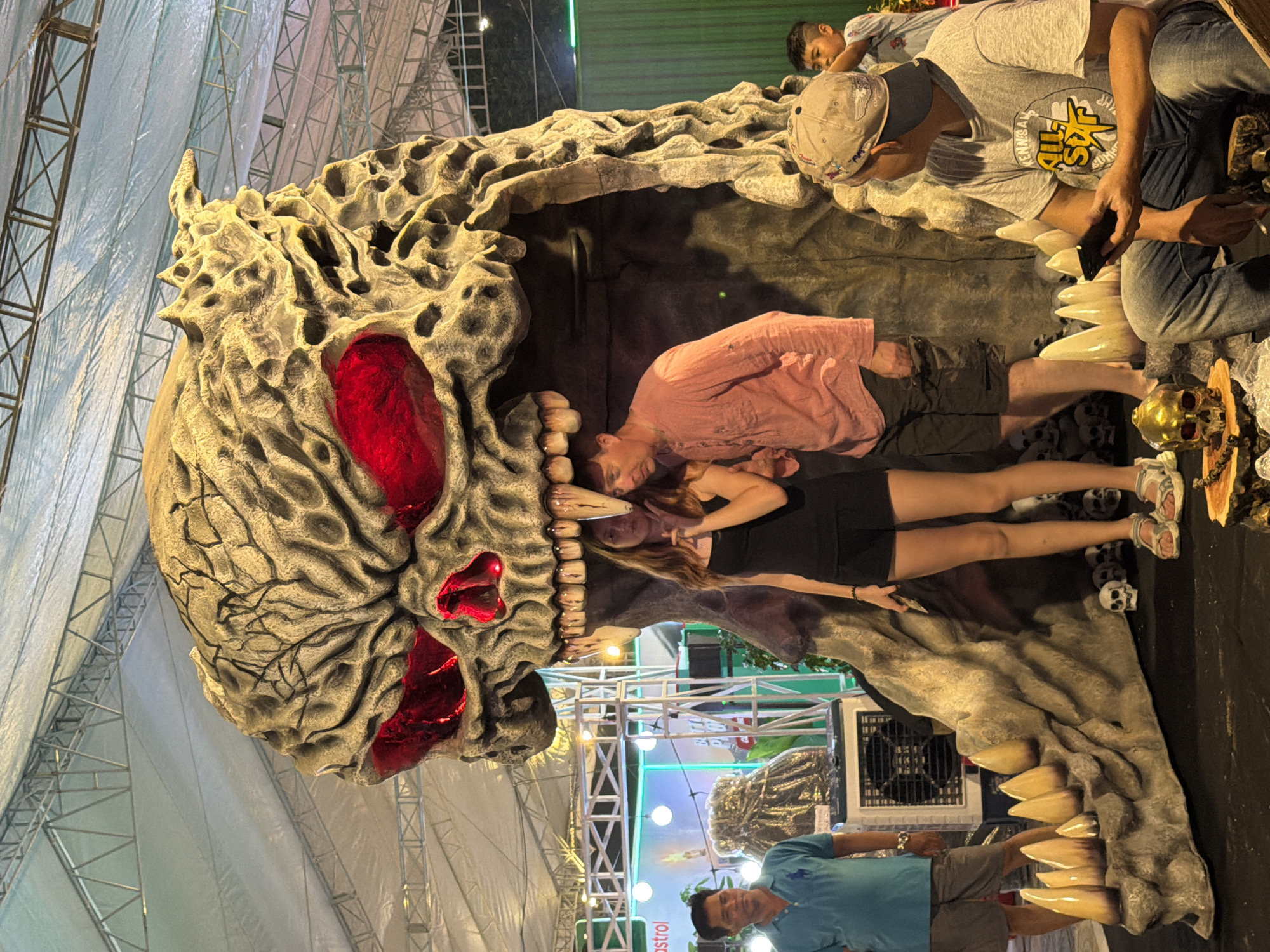 The image shows a large skull-shaped structure with people posing inside its mouth. The skull has red, cavernous eye sockets and large, pointed teeth. People are standing and sitting inside the mouth for photos, with others visible around the structure. The background includes scaffolding, lighting fixtures, and what appears to be a promotional display. The image suggests a temporary installation or attraction, possibly related to Halloween or a themed event. The location is identified as Nha Trang Ward, Vietnam.