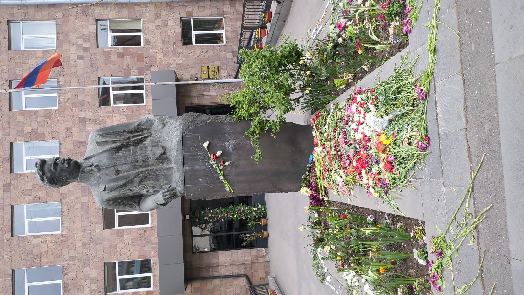 The image displays a bronze bust of a man, seemingly a memorial, situated outdoors against a large, multi-story building. The statue, depicting the man from the waist up in a suit jacket and collared shirt with a somber expression, rests on a tall, dark, rough-hewn stone base.
Abundant fresh and slightly wilted flowers, including various colorful blossoms like carnations, gerberas, and others, are piled in a large mound in a planter directly beside the statue's base. Another bouquet, consisting mainly of white calla lilies and red roses, is carefully laid upon the dark stone base itself.
The backdrop is a building with a light brown, possibly granite or brick, facade featuring numerous rectangular windows with white frames. The national flag of Armenia (red, blue, orange horizontal stripes) is prominently flying on a pole to the left of the building. Green shrubbery and smaller plants are visible in garden beds around the memorial, and the ground is paved with dark grey stones or asphalt.
The scene strongly suggests an act of remembrance, a tribute, or a memorial event. The lighting indicates daytime with soft, even illumination. The Armenian flag confirms the location as Yerevan, Armenia. Notable visible text includes a cursive signature, likely in Armenian script, inscribed on the dark stone base of the statue, possibly "Hovhannes Tumanyan". A small golden plaque is affixed to the building wall but its text is illegible. The overall atmosphere conveyed is one of respect and solemnity.
