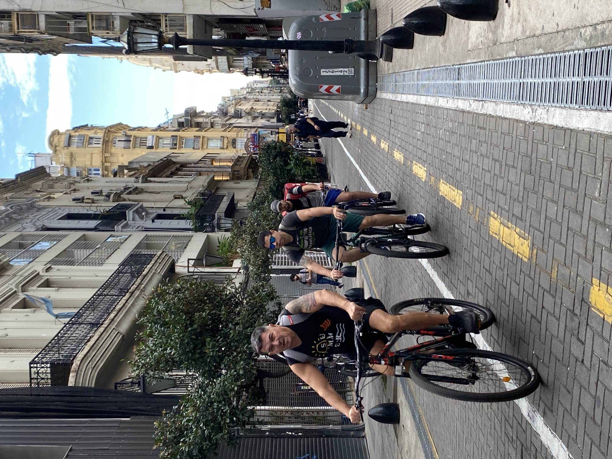 The image shows a group of three men riding bicycles on a street in Buenos Aires, Argentina. The street is paved with grey bricks and marked with yellow lines indicating a bike lane. A metal grate runs along the edge of the bike lane. A trash receptacle is situated on the side of the street. Buildings of varying heights line the street, with architectural details such as balconies, windows, and fire escapes visible. An Argentinian flag is visible on one of the buildings. People can be seen walking in the background. The sky is partially cloudy. The men are riding towards the camera. The first man has tattoos on his arm and is wearing a black shirt with a logo. The second man is wearing sunglasses and a black shirt. The third man is wearing a blue shirt. Each bicycle is equipped with a front light.