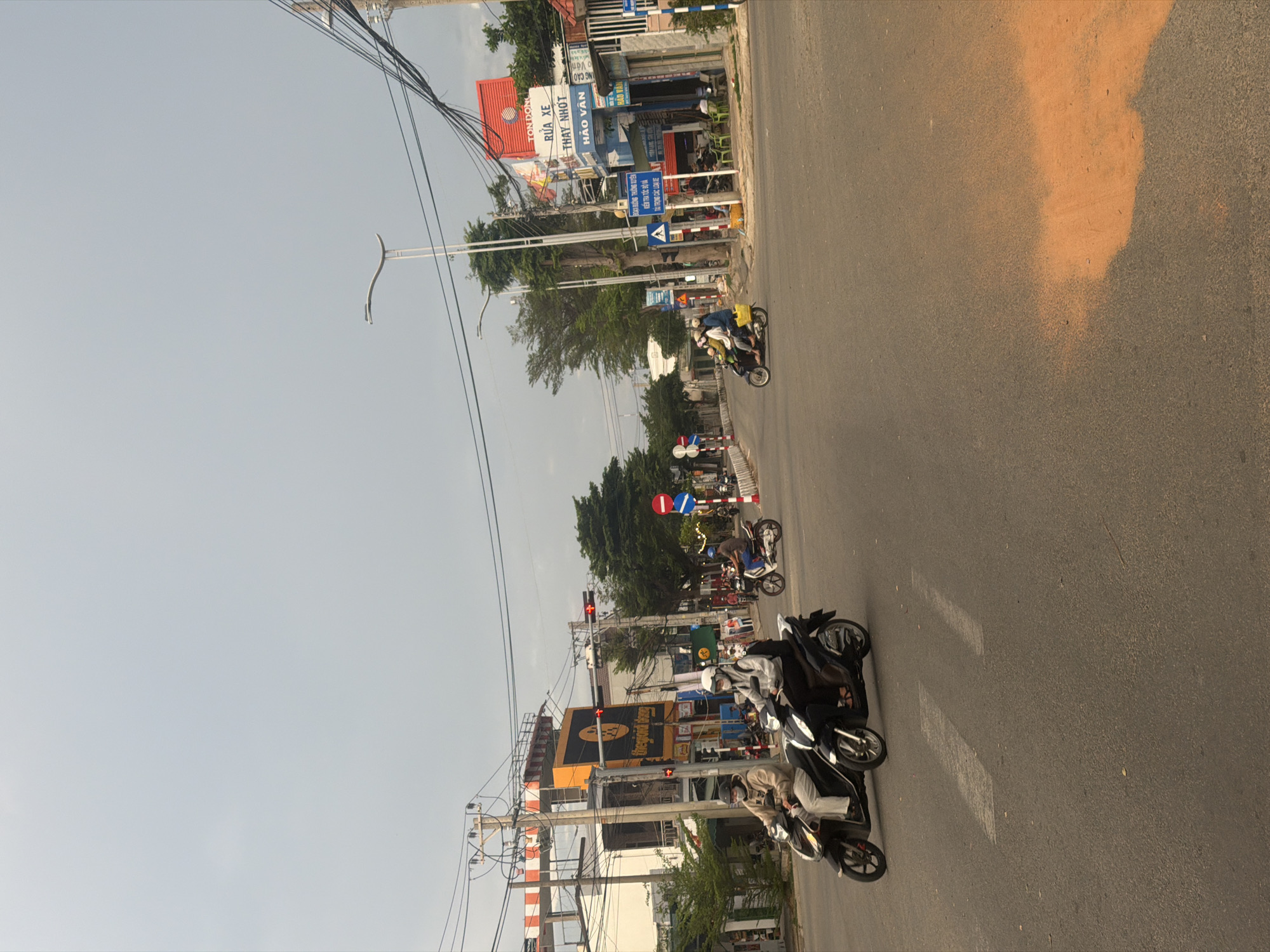 The image depicts a street scene in Dong Hai Ward, Vietnam, captured in an upright portrait orientation despite the provided file being rotated. The photograph shows a bustling road with several motorbikes, buildings, and overhead utility lines under a clear, light sky, likely during late afternoon or early morning.
In the foreground, two individuals on black motorbikes are visible, both wearing helmets, seemingly waiting at a red traffic light. The asphalt road they are on features faint white markings. To the right of the road, a large, irregularly shaped reddish-brown area contrasts sharply with the dark grey pavement.
Further down the road, more motorbikes and pedestrians can be seen, with a few other motorbikes moving or parked. A prominent yellow building with a dark logo, possibly resembling "thegioididong" or a similar commercial establishment, stands out. Buildings line both sides of the street, displaying various commercial signs. Several utility poles with a dense network of overhead wires crisscross the sky.
Visible text on signs includes "thegioididong," a blue sign warning "ĐOẠN ĐƯỜNG THƯỜNG XUYÊN KIỂM TRA TỐC ĐỘ VÀ PHẠT NẶNG CÁC LOẠI XE" (Stretch of road frequently checked for speed and heavy fines for all types of vehicles), and a red sign advertising "RỬA XE THAY NHỚT" (Car Wash, Oil Change). Other shop names like "HẢO VÂN" and "TÔN ĐÔNG" are also discernible. Traffic lights are illuminated red, indicating a stop for vehicles. The overall scene captures a typical moment of urban daily life and traffic in Vietnam.