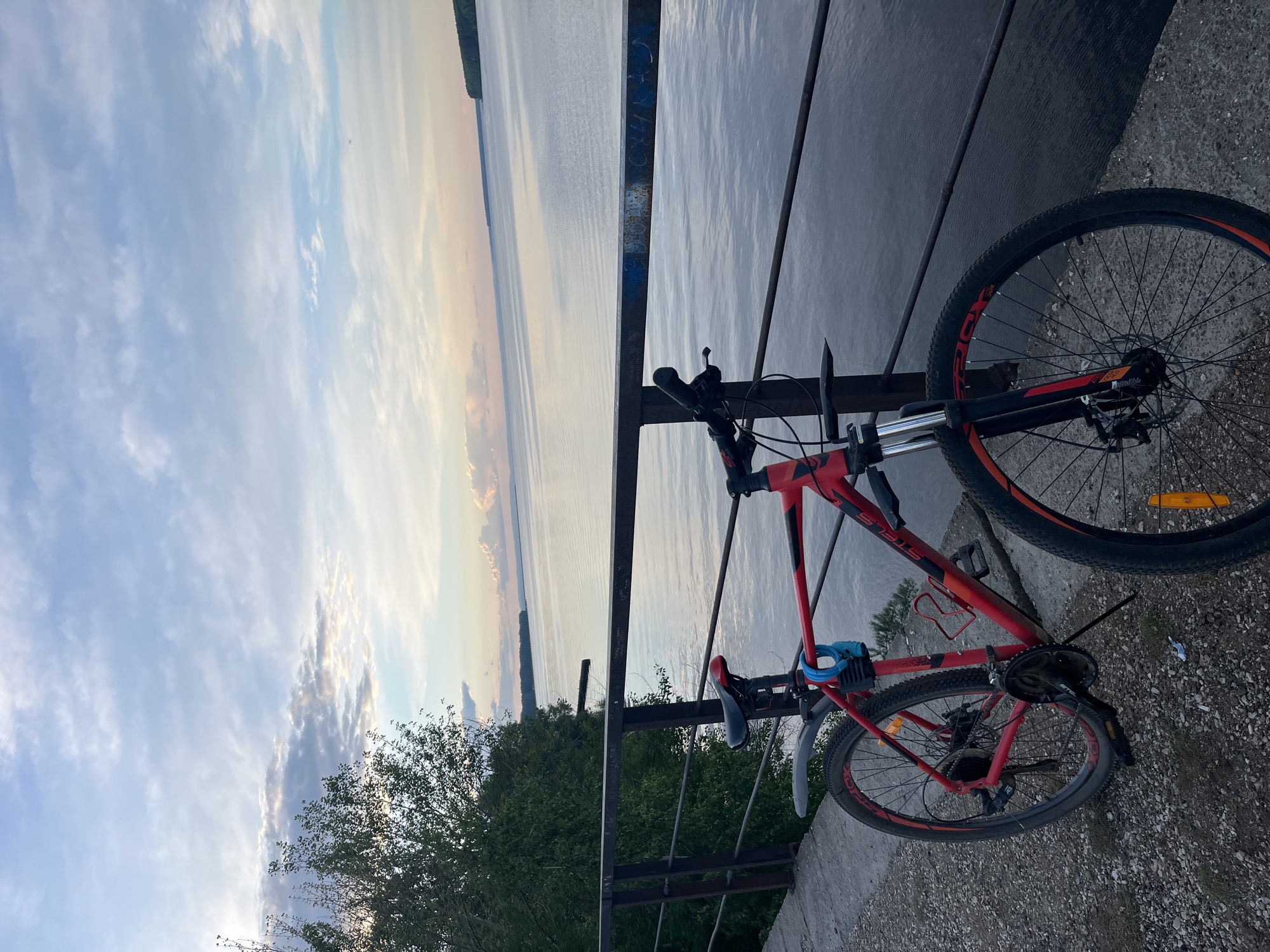 A red bicycle is leaning against a railing overlooking a large body of water, likely a river or reservoir. The sky is cloudy with hues of blue, gray, and a touch of orange at the horizon, suggesting either sunrise or sunset. The railing is metal and appears to be slightly rusted. The bicycle is parked on a concrete surface, possibly a bridge or a viewing platform. The presence of a tree suggests the location might be a park or a landscaped area. Given the city mentioned, this is likely the Votkinsk Reservoir on the Kama River in Chaikovsky, Russia.