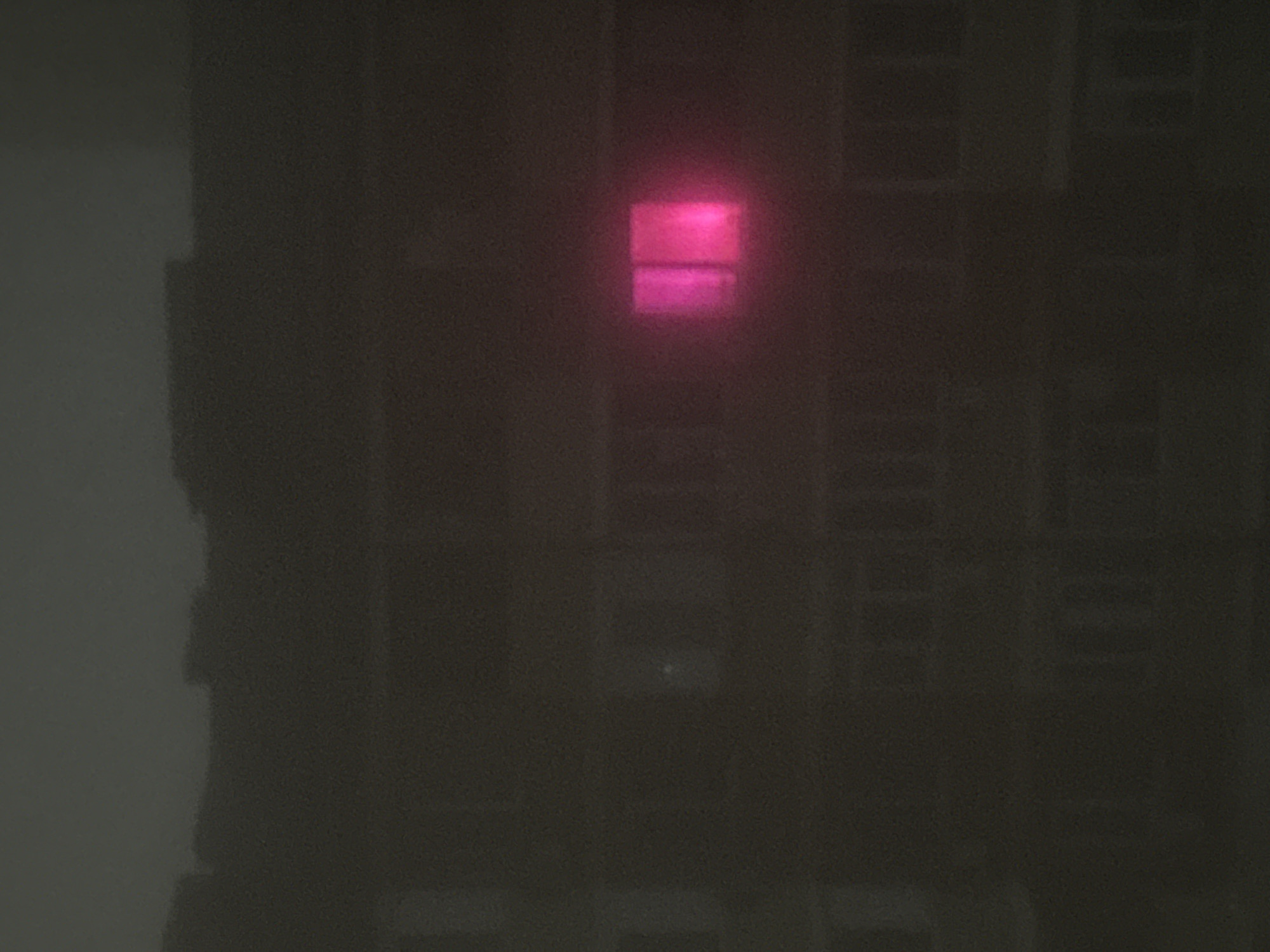 The image shows the exterior of a dark building at night. A single window emits a bright pink light, contrasting sharply with the surrounding darkness. The rest of the building's windows are visible as dark, indistinct shapes. The left side of the image appears to be a lighter-colored wall adjacent to the building. The location is Unknown, Unknown.