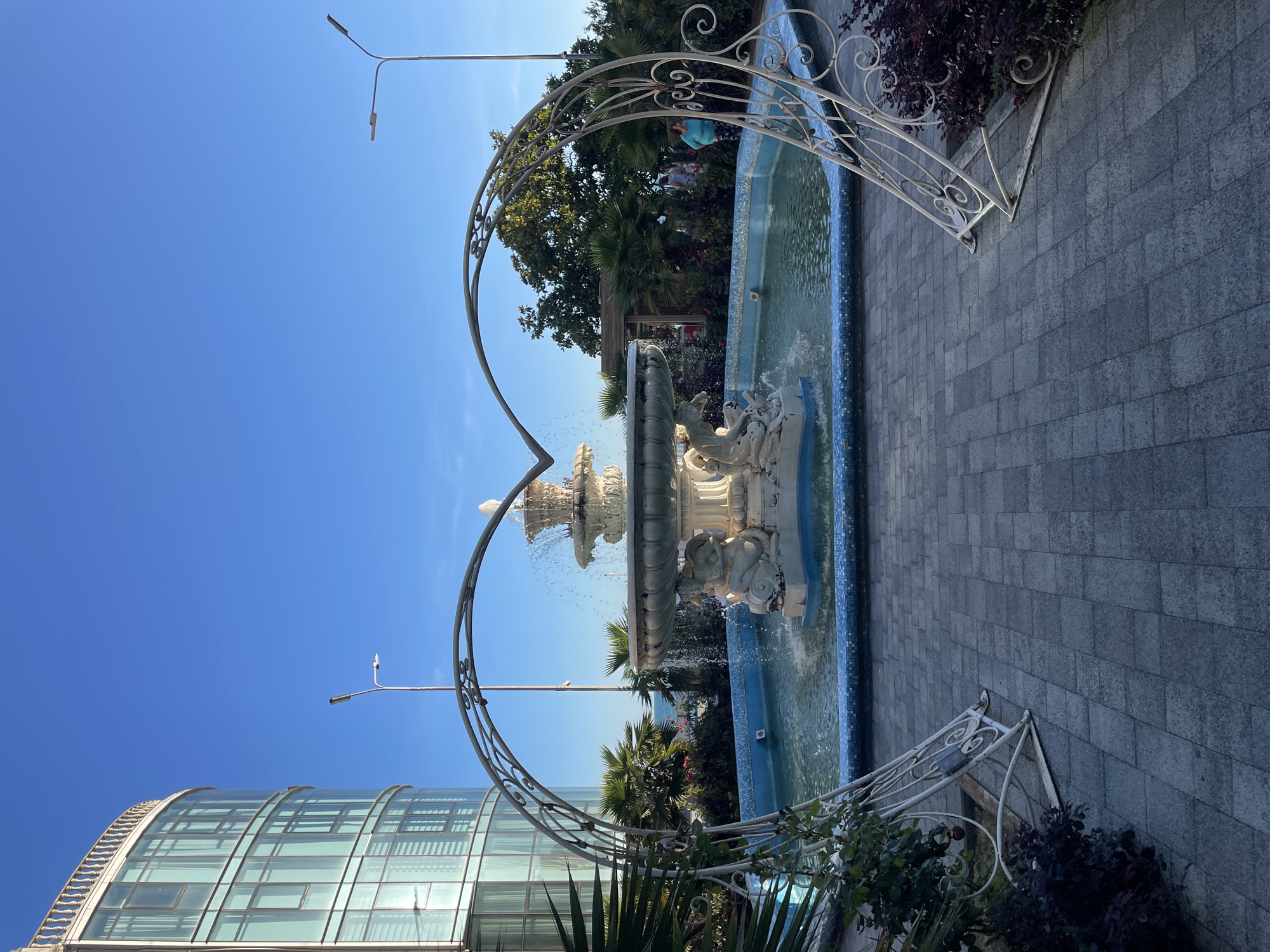 This is a photograph taken in Sochi, Russia, depicting an outdoor scene with a fountain as the central subject. The fountain is ornate, with multiple tiers and sculpted figures, and water is actively spraying from it. The fountain is situated within a rectangular pool lined with blue tiles.
In the foreground, a decorative white metal archway, shaped like a heart, frames the fountain and the pool. This archway is adorned with scrolling metalwork. To the left of the fountain, a modern building with a curved glass facade is visible, reflecting the clear blue sky. To the right and behind the fountain, lush green foliage, including palm trees, adds to the setting. A few people are visible in the background near some bushes, suggesting it's a public area.
The paving around the fountain consists of large, grey rectangular tiles. A lamppost is visible in the upper left portion of the image, and another in the upper right. The bright blue sky suggests it is a sunny day. The time of day appears to be daytime, possibly late morning or afternoon, given the clear sunlight. The overall atmosphere is pleasant and likely a tourist or recreational spot. There is no visible text in the image.