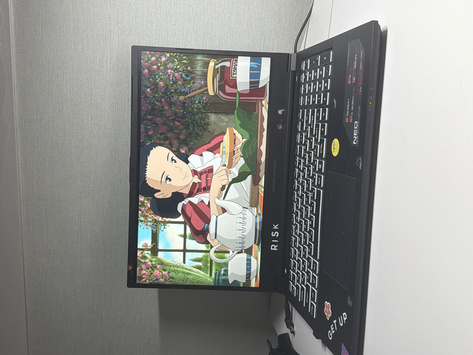 The image shows a black laptop with a screen displaying an animated scene of a young woman preparing food. The laptop is open and positioned on a white surface, likely a desk, with a light gray textured wall in the background. The animated scene features the woman in an apron, in front of a window with floral curtains, and a table with a teapot, a jar of jam, and a plate of food. The laptop has a keyboard with white lettering and a trackpad. There are several stickers on the laptop's palm rest, including one that says "GET UP" and another with a flower. The overall setting appears to be an indoor space, possibly a room in a home or office. There is no indication of the specific location within Dongducheon-si, South Korea, as no recognizable landmarks or context clues are present.
