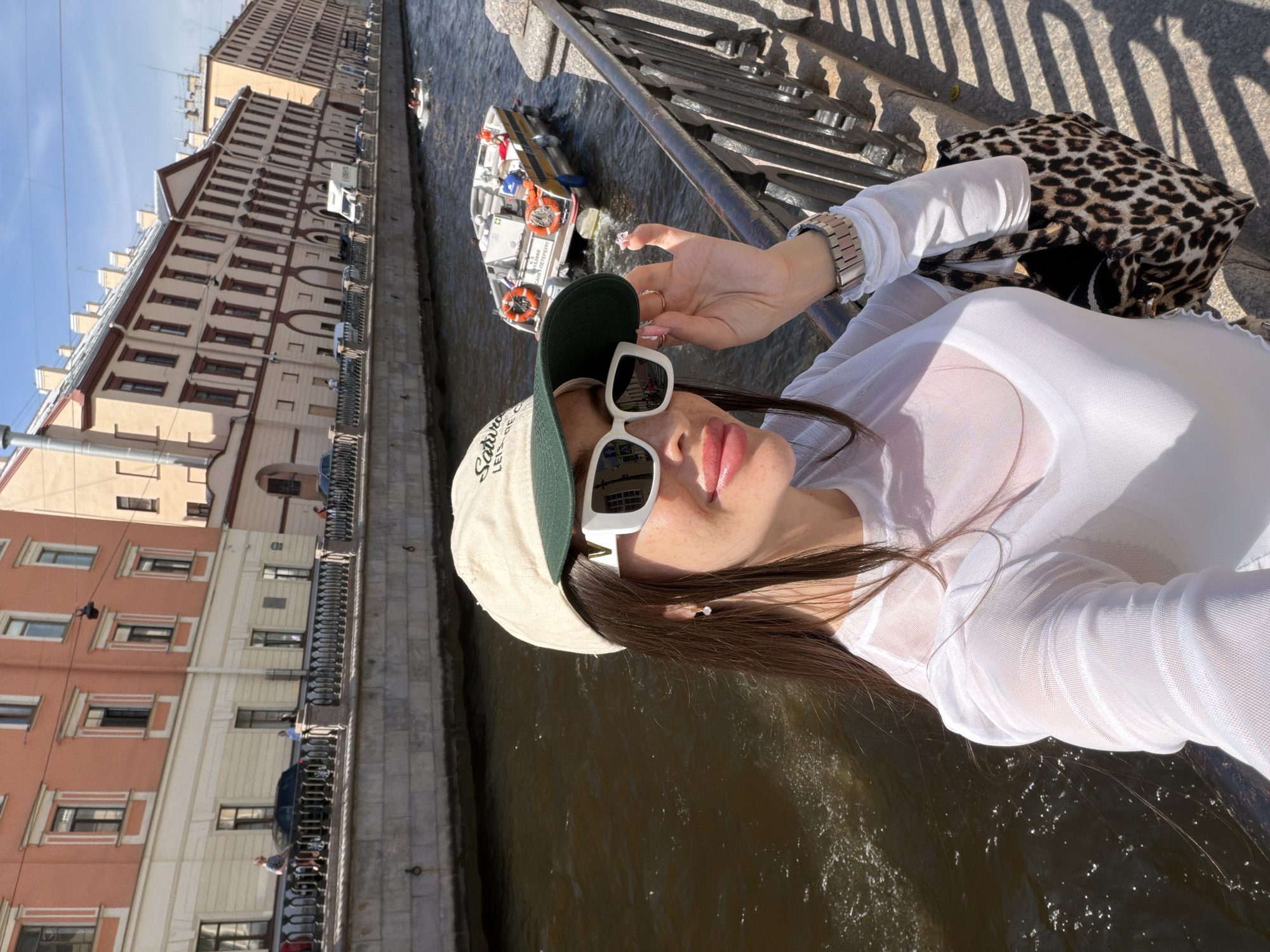 The image shows a woman taking a selfie on a sunny day in Saint Petersburg, Russia. She is standing on a canal-side embankment, with buildings and a boat visible in the background. The woman is wearing a white sheer long-sleeved shirt, a beige baseball cap with a green brim, and white-rimmed sunglasses. She is holding her sunglasses with her right hand. A leopard-print bag is slung over her left arm. The embankment is paved with stone, and a black metal fence runs along the edge. In the canal, a white and yellow boat with several people on board is moving away from the viewer. The buildings in the background are tall and multi-story, with traditional architectural features. The sky is blue with scattered white clouds.