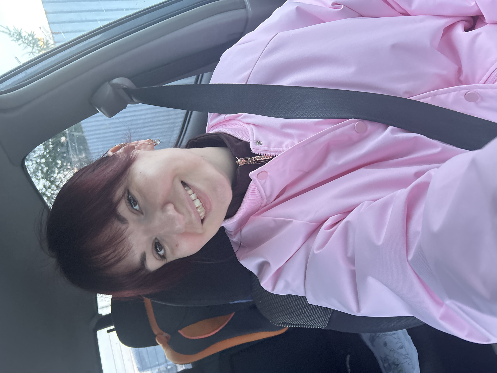 A woman is sitting in the driver's seat of a car, wearing a pink jacket and a seatbelt. A child's car seat is visible behind her. The car is parked in Фоки, Russia.