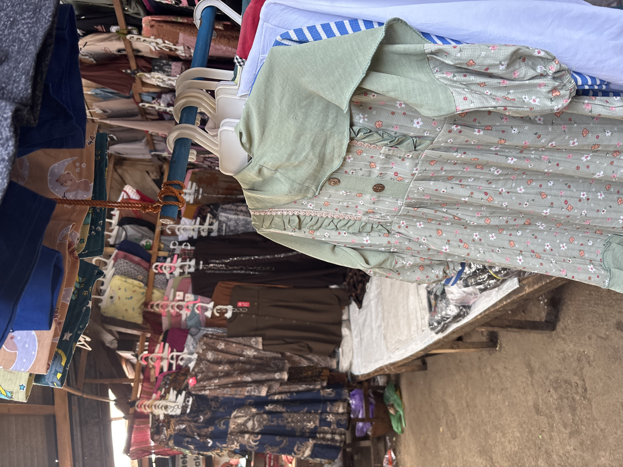 The image depicts an outdoor market stall in Tirtomoyo, Indonesia. The stall displays a variety of clothing items, primarily dresses and shirts, hanging from a blue metal rod. In the foreground, a light green floral-patterned dress with ruffled detailing and balloon sleeves is prominently featured. Behind it, other garments in various colors and patterns are visible, including a dark brown dress, a navy blue patterned dress, and a white shirt with blue stripes. The stall is set up along a wooden bench covered with a white cloth, upon which some clothing items are also placed. The ground appears to be dirt and gravel, with some plastic bags and discarded items scattered around. The market seems to be covered by a makeshift roof structure made of corrugated metal and fabric. The overall impression is one of a bustling, informal marketplace.