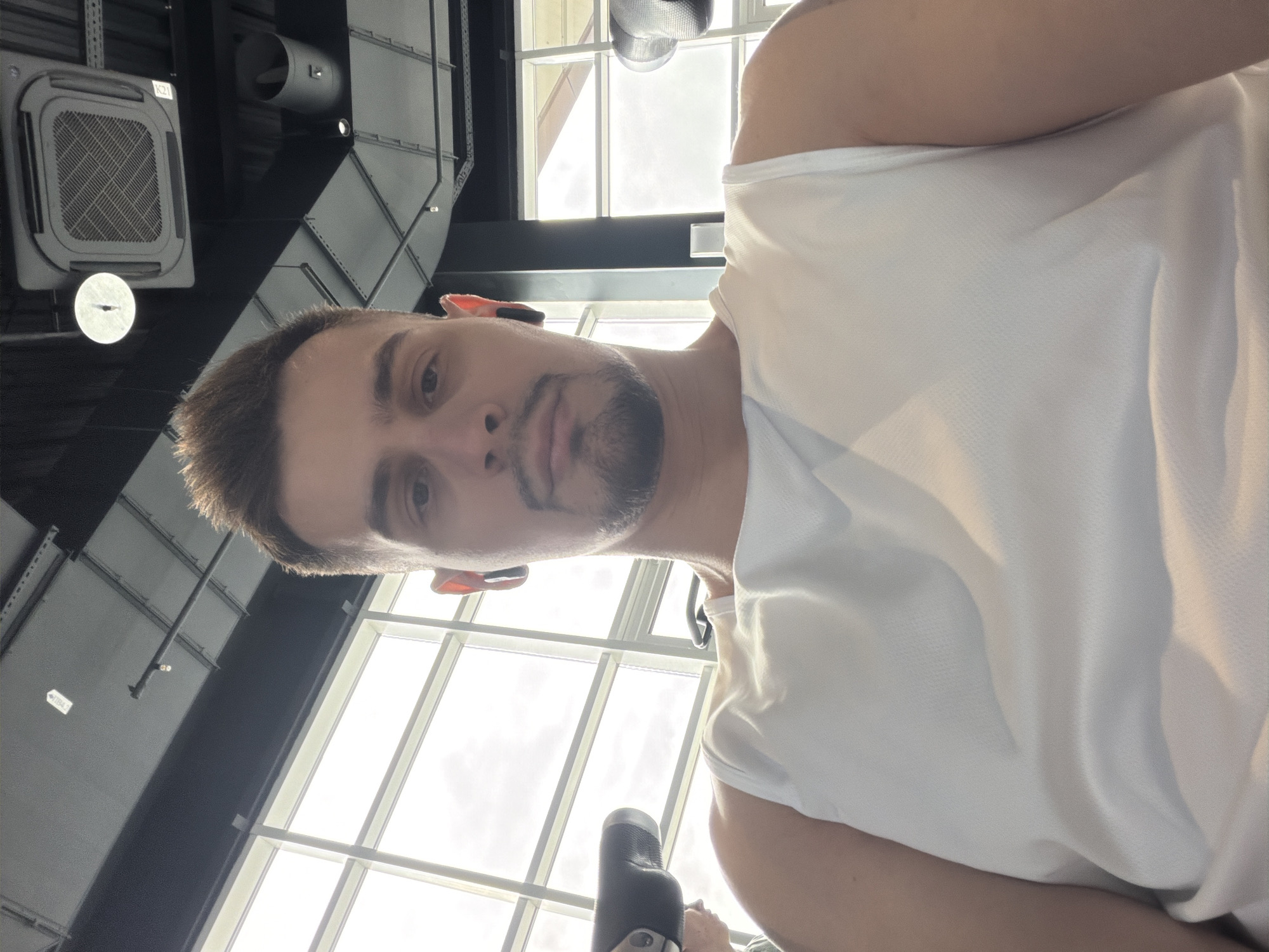 A low-angle selfie shows a man with short brown hair, a trimmed beard, and fair skin, wearing a white tank top. He is facing the camera with a neutral expression. Earbuds are visible in both ears. The background features a large window with a grid pattern, allowing a view of the sky outside. Structural elements of the ceiling and lighting fixtures are also visible. The location is not determinable from the image.