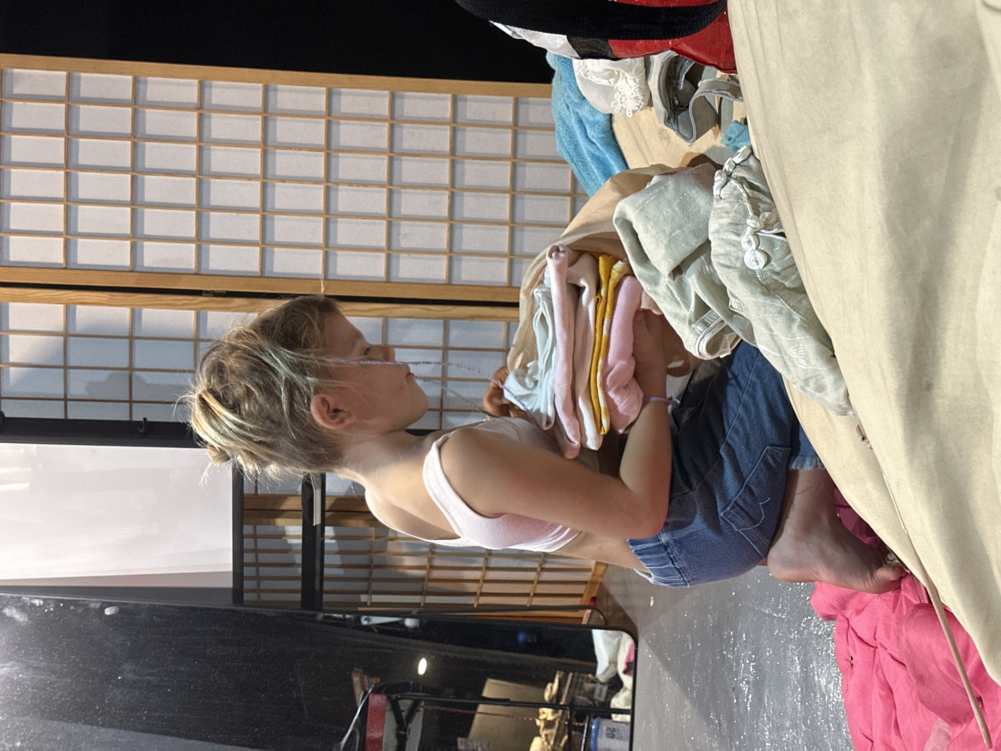 An indoor scene features a young child, possibly a girl, with light-colored hair pulled back, sitting or kneeling on a dark, somewhat reflective floor. She is wearing a light pink sports bra or crop top and blue denim shorts or overalls, and is barefoot. The child is holding a neatly folded stack of colorful clothes in shades of pink, yellow, light blue, and white, appearing focused on the task of sorting or organizing them. A small light purple bracelet adorns her right wrist.
In the background, a large, light-colored Japanese-style shoji screen or room divider with a grid pattern of wooden frames and translucent panels provides a structured backdrop. To the child's right, a large piece of beige or tan fabric drapes down, partially covering a pile of light green and white clothes. A bright pink fabric is also visible on the floor in this area. The lighting is bright and even, illuminating the domestic activity. No visible text is present in the image. The setting is a casual indoor environment in ตำบลเกาะพะงัน, Thailand.