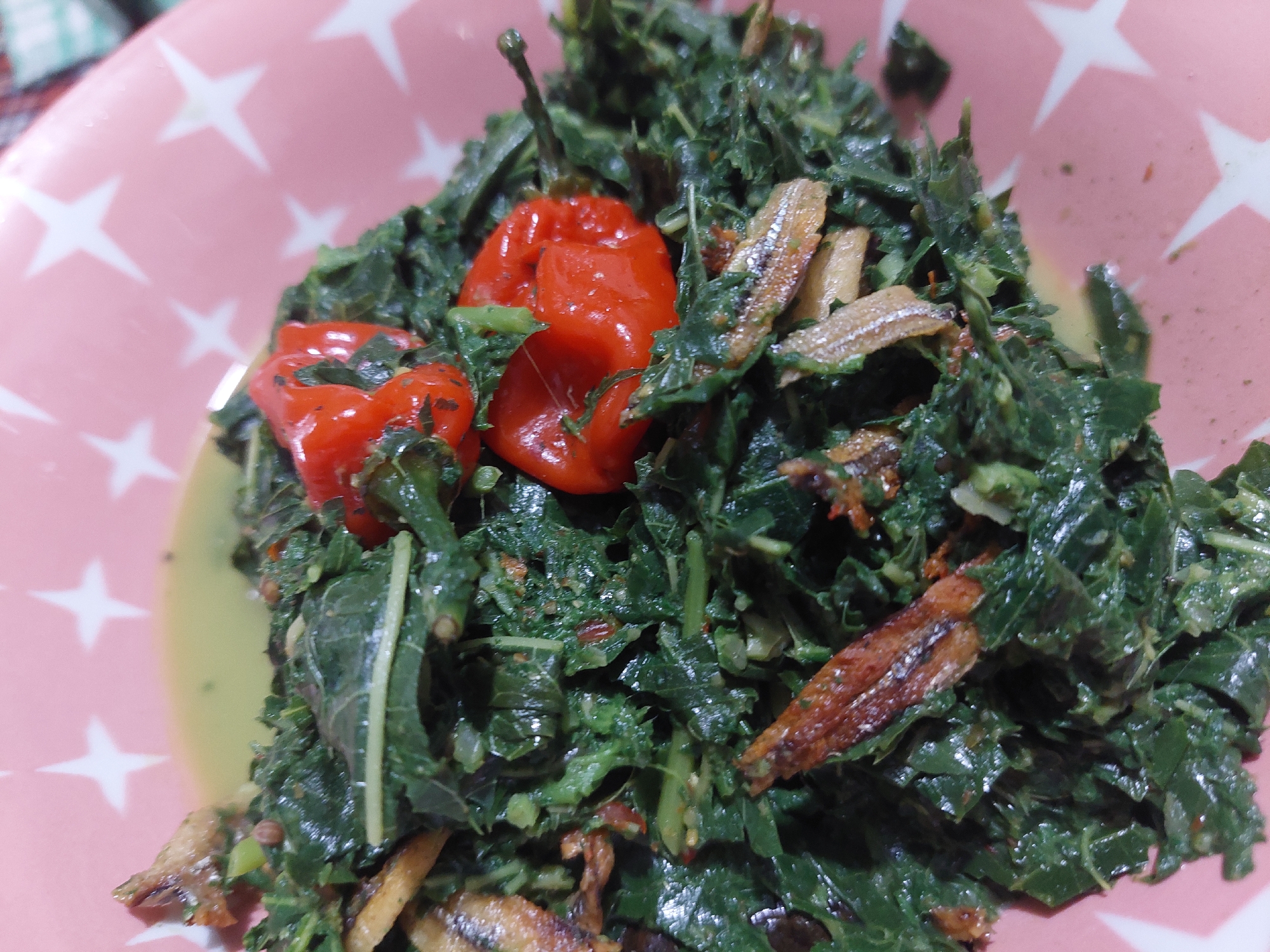 A close-up, overhead shot shows a pink plate with white stars, filled with a dish of cooked green leafy vegetables and small, fried fish. Two whole red chilies are visible in the center of the dish. The food appears to be in a light sauce. The lighting suggests an indoor setting. The dish is likely a local Indonesian cuisine, possibly from the Jelutung area in Indonesia.