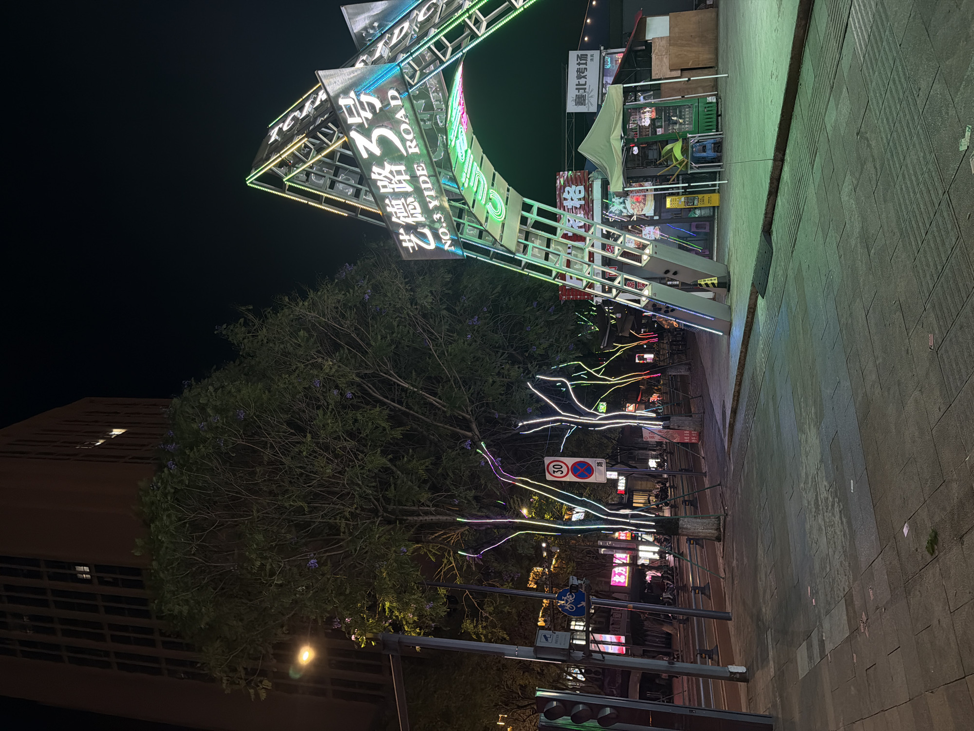 The image captures a night scene in Chongqing High-tech Zone, China. At the forefront is a large, illuminated archway structure with Chinese and English signage, including "NO.3 YIDE ROAD." A building sits to the left. The walkway features decorative lights strung through the trees. Seating for an outdoor eatery is visible. A traffic sign indicating a speed limit of 30 is positioned along the roadway.