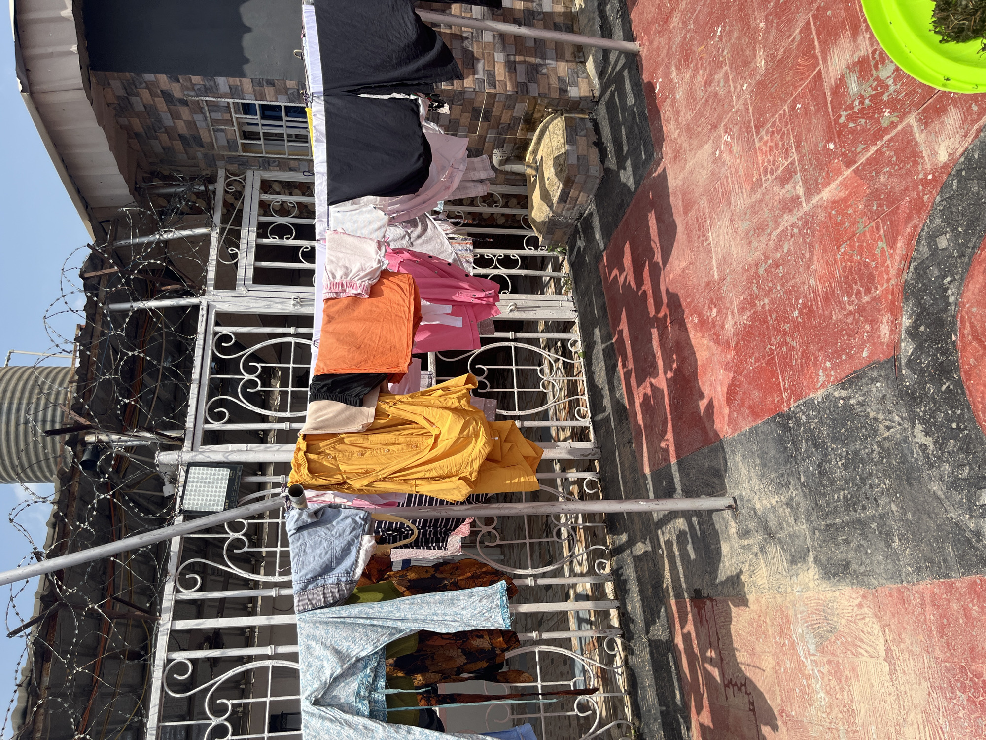 In Abuja, Nigeria, this outdoor scene depicts a clothesline hung with various items of laundry. The clothes are draped over a metal line supported by upright posts, with a decorative white metal fence visible in the background. Above the fence, barbed wire is strung along the top, suggesting a security measure. A large, grey water tank is positioned on the roof of a building behind the fence, and part of a brick-patterned wall is visible to the right. The ground is paved with red and black tiles, showing signs of wear and some dust accumulation. Sunlight casts shadows from the clothesline and laundry onto the tiled surface.