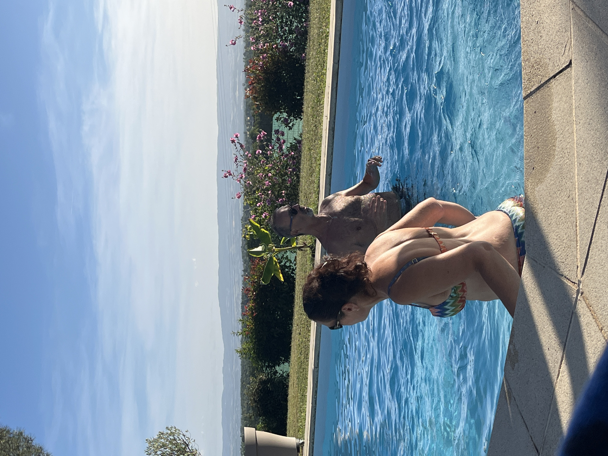 Two individuals are pictured in a swimming pool with clear blue water, set against a backdrop of lush greenery and a distant landscape. The woman, in the foreground and facing away from the camera, wears a colorful bikini. The man, partially submerged and facing the woman, has his back to the camera. The scene is bathed in sunlight under a bright blue sky with scattered clouds. The swimming pool area appears to be in a private residence, likely in the vicinity of Saint-Galmier, France, given the typical landscape of the region. The surrounding environment features manicured lawns, flowering bushes, and mature trees, suggesting a well-maintained outdoor space.