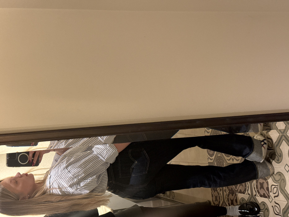 The image displays an indoor scene in Tel-Aviv, Israel, captured as a mirror selfie. The primary subject is a young adult female with long blonde hair, seen from the mid-thigh up, angled slightly to her right and looking into a large mirror over her left shoulder. She is dressed in a light-colored, blue-striped, collared shirt with rolled-up sleeves and dark blue denim Levi's brand jeans featuring distinct arcuate stitching on the back pockets. Her right hand holds a dark-colored smartphone to take the photograph. She is wearing light-colored athletic shoes. To the far left of the frame, reflected in the mirror, is the lower leg and foot of another individual, attired in dark pants, white socks, and black patent leather loafers. The floor is covered with ornate, patterned tiles in a palette of beige, grey, and dark brown. The visible wall surfaces are plain and light beige. Artificial lighting illuminates the area.