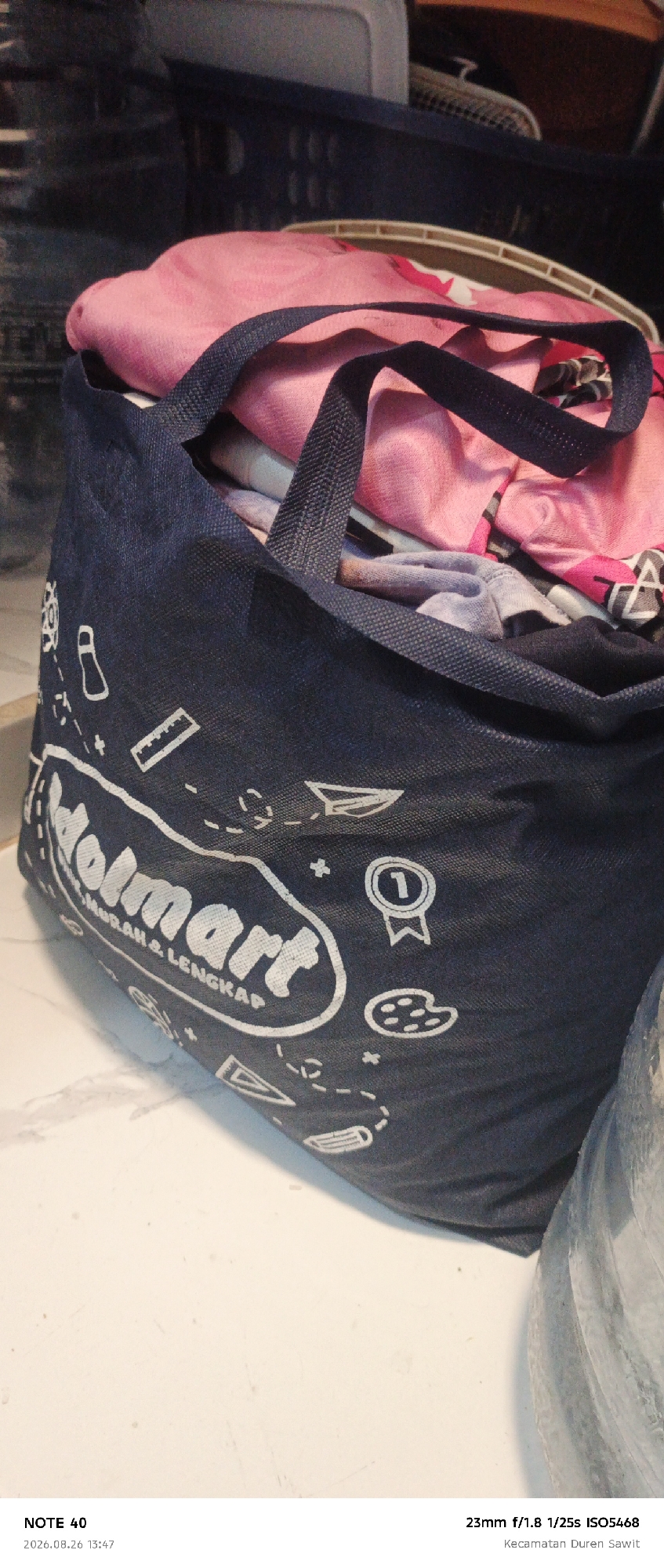 The image is a close-up shot of a dark blue reusable shopping bag filled with pink and grey clothing items. The bag has white graphics, including the text "olmrt MURAH & LENGKAP" and various icons such as a trophy, a palette, and a paper airplane. A portion of a clear plastic bottle is visible on the right side of the frame. The bag rests on a light-colored, speckled surface that appears to be a countertop or table. The background is blurred and shows dark blue laundry baskets and other indistinct household items. The timestamp "2026.08.26 13:47" and camera details "23mm f/1.8 1/25s ISO5468" along with "Kecamatan Duren Sawit" are present at the bottom of the image. The location specified, Kecamatan Duren Sawit, is an administrative district within East Jakarta, Indonesia.