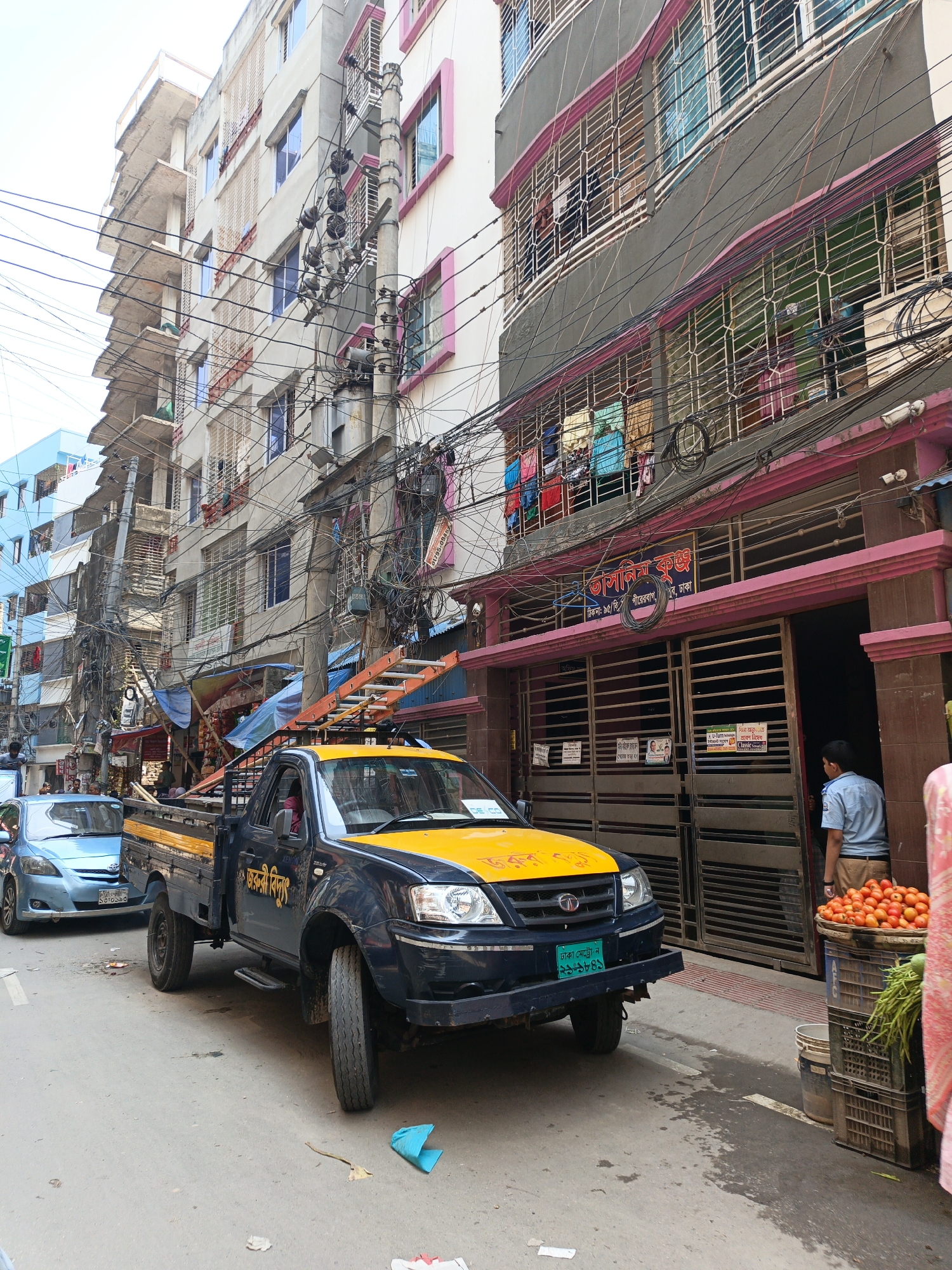 The image captures a bustling street scene in Dhaka, Bangladesh. In the foreground, a black and yellow utility truck is parked on the side of the road, bearing the license plate "ঢাকা মেট্রো-ন ২১-১৮৪১". It has the text "জরুরী বদ্যুৎ" (Emergency Electricity) printed on it, indicating its purpose. A ladder is placed on top of the truck. Behind the truck is a light blue car.
Adjacent to the truck, on the right, is a building with pink trim and a gray facade. A large metal gate is visible at the entrance, with a sign that reads "তাসনিম কুঞ্জ", followed by "ঠিকানা: ৯৫/ কি পীরেরবাগ, পুর, ঢাকা". Baskets of tomatoes and vegetables are positioned by the entrance.
The buildings in the background are multi-storied with various paint schemes. Tangled electrical wires crisscross above the street. People are visible walking and standing in the background, some of them operating stalls or shops. Overall, the image depicts a typical urban street scene in Dhaka, with its unique blend of infrastructure, commerce, and dense population.