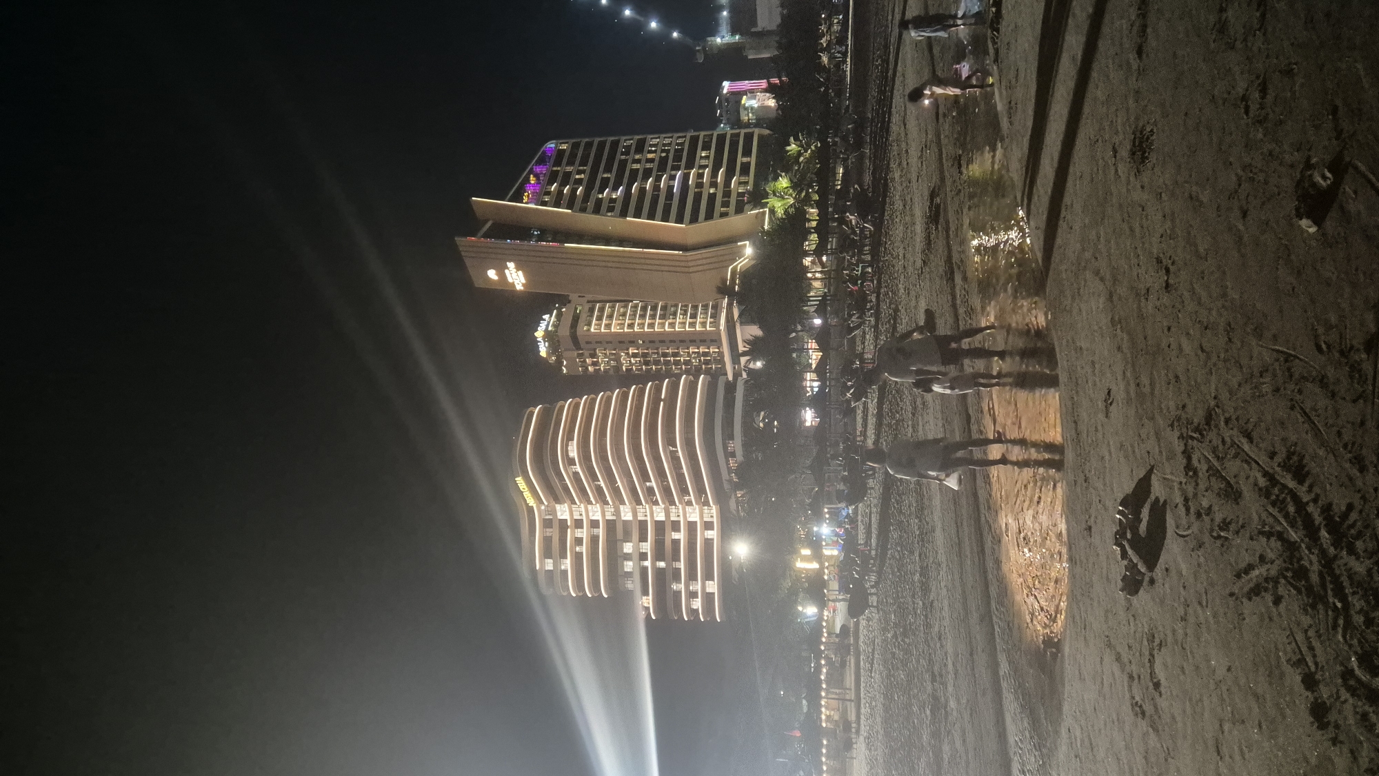 The image captures a nighttime scene on a sandy beach in Da Nang, Vietnam, with several illuminated buildings in the background. In the foreground, a family of three, a man, a woman, and a child, are walking through shallow water on the beach. The man and woman appear to be holding hands, and the child is walking between them. Their reflections are visible in the water. In the distance, there are rows of beach chairs and umbrellas. The buildings in the background are modern high-rises, with the "VISTA CELLA" building prominently featured. The lighting from the buildings casts a warm glow on the scene. The sky is dark, indicating it is nighttime.