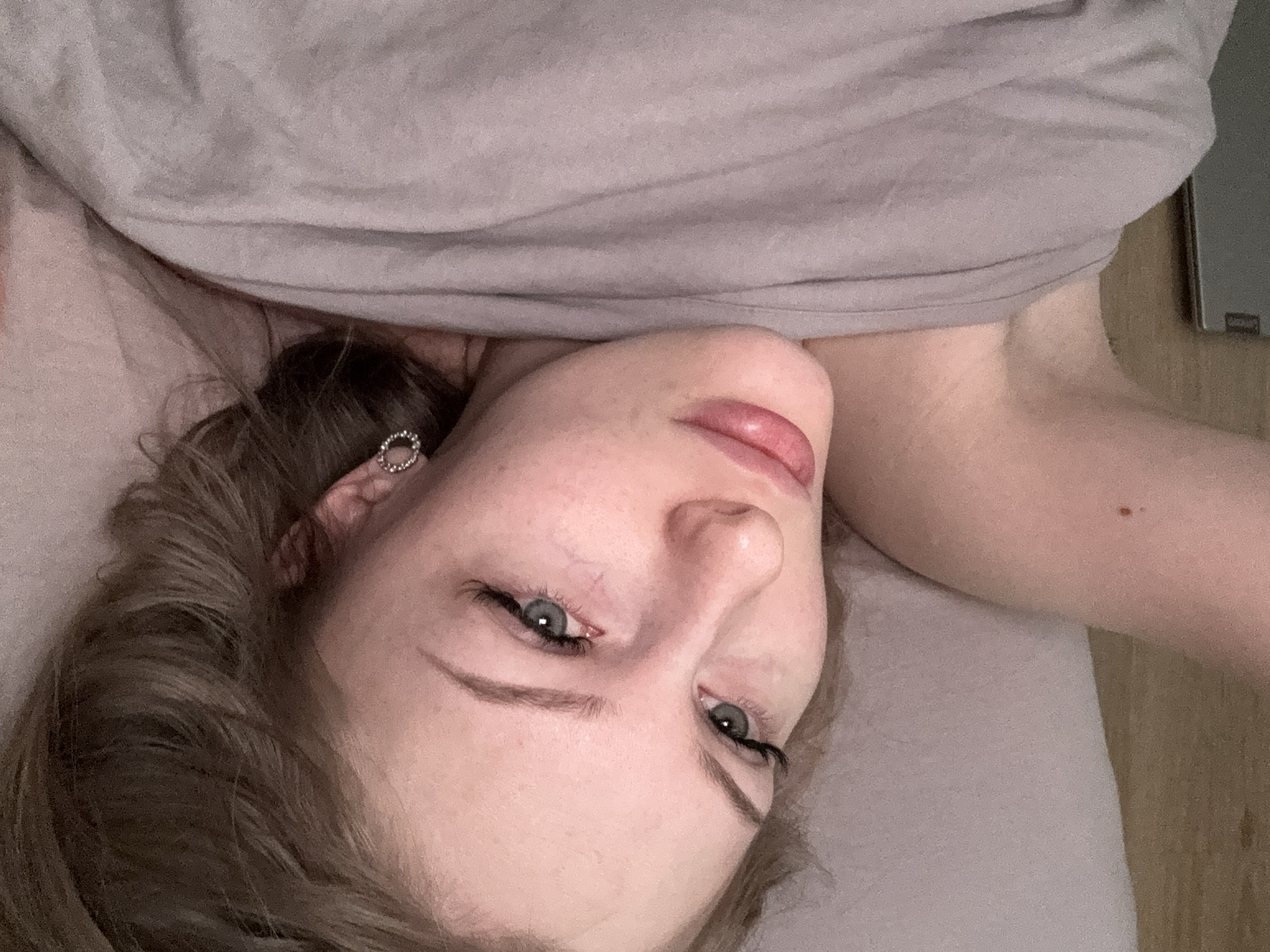 A light-skinned person with light brown hair lies on a light gray surface, possibly a bed or a similar soft surface. The person is angled toward the viewer, with their head tilted. The person has light gray eyes and is wearing what appears to be an earring in their left ear. A gray fabric is positioned above the person's head. The environment has light-colored wooden flooring visible, and part of a laptop is visible in the upper right corner. There is no visual information to suggest the location is in Perm, Russia.