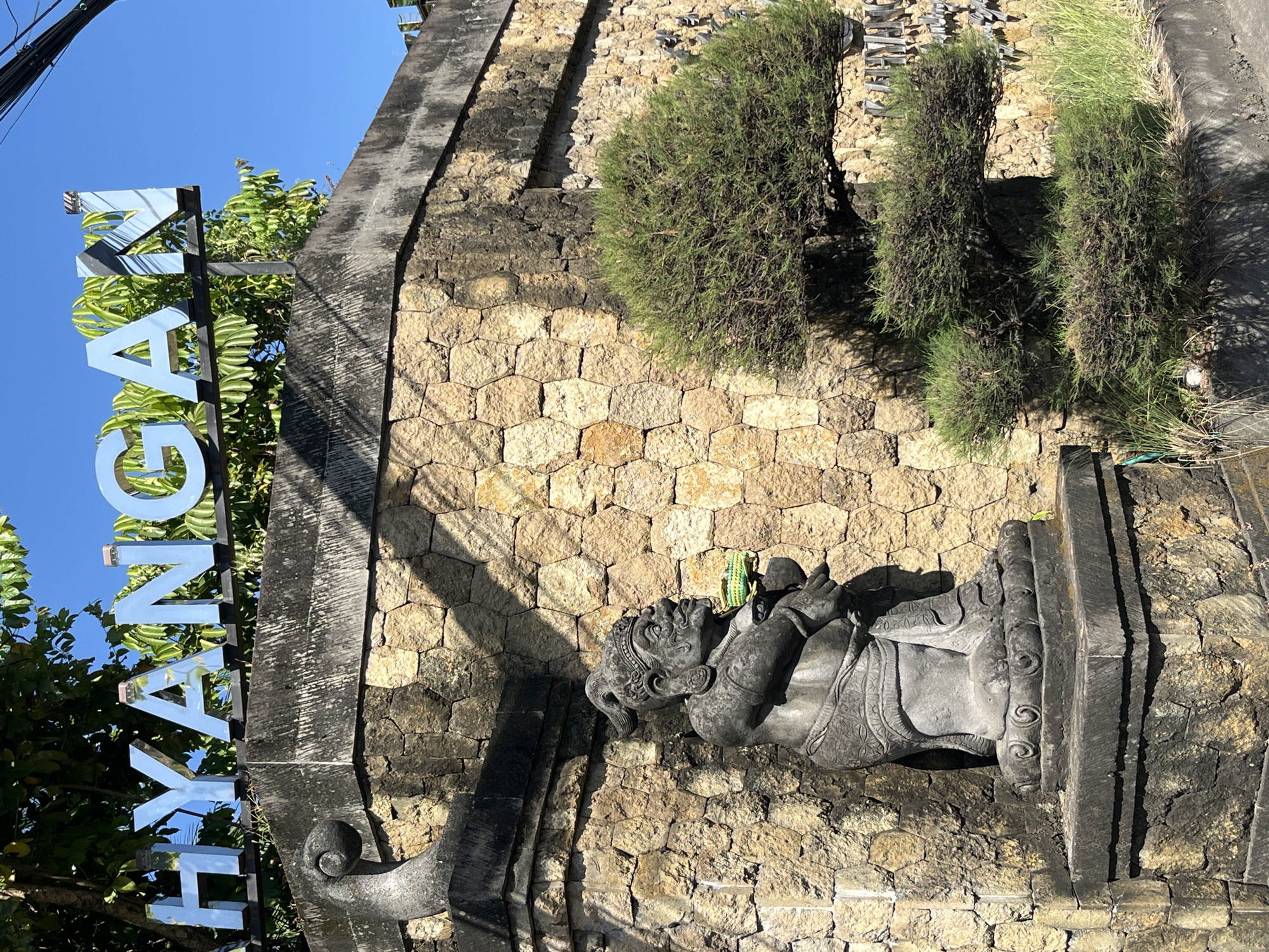 The image shows a stone statue of a seated figure, holding a green object, in front of a stone wall. Above the wall, there is a sign with the word "HYANGAN" in large, silver, reflective letters. A small, manicured tree is positioned to the right of the statue, in front of the wall. The wall itself is constructed from irregularly shaped stones and shows some signs of weathering. Sunlight casts shadows on the wall and statue, indicating it is daytime. The statue is dressed in traditional attire. The location appears to be an entrance to a place called "Hyangan" in Sukawati, Indonesia, suggested by the sign and the traditional Balinese-style sculpture.