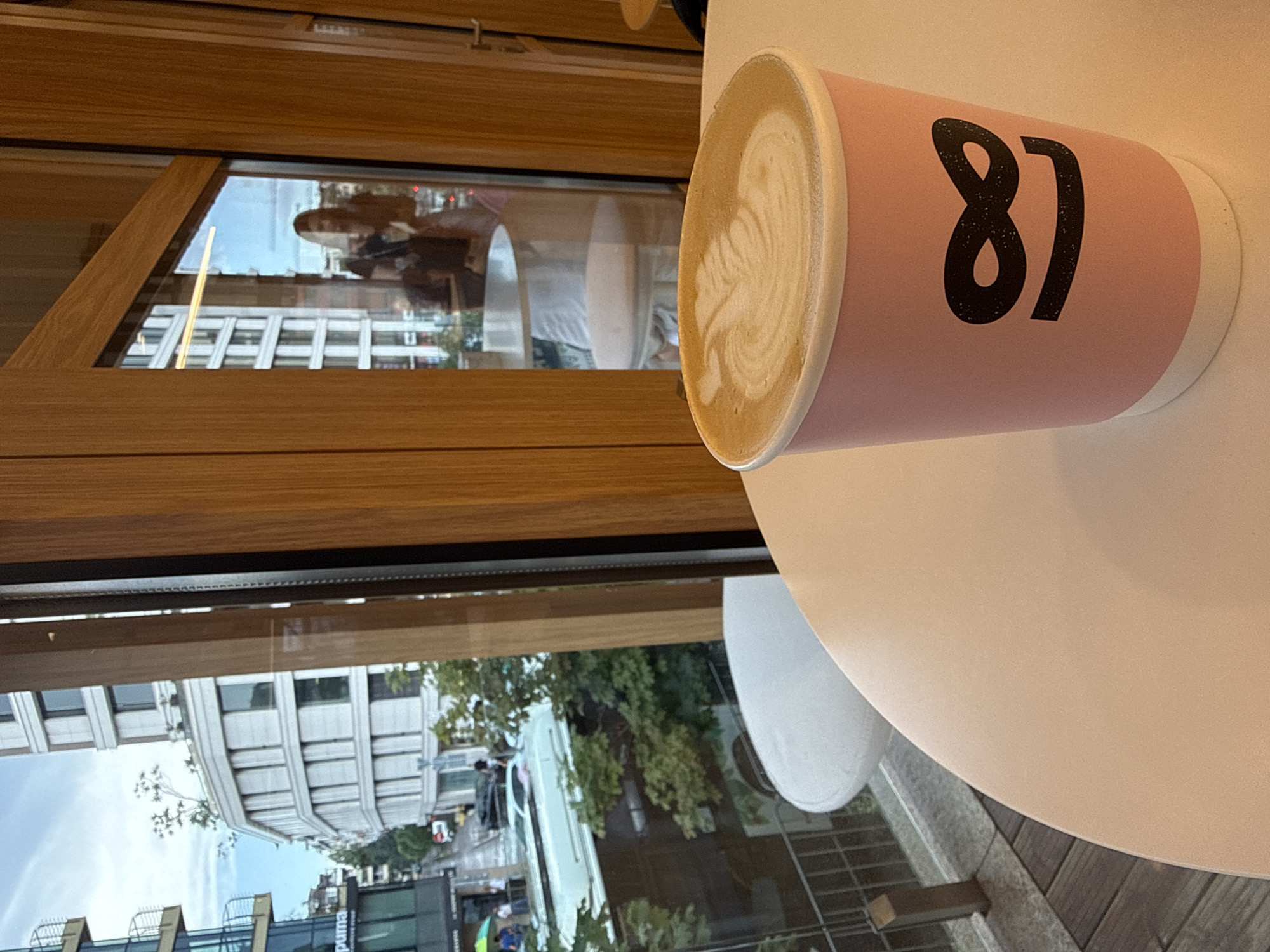 In the foreground of the image, a light pink paper cup with a white base is placed on a white circular table. The cup features a black graphic of the number "81" stylized to resemble a smiling face. The coffee inside the cup has latte art with a fern-like pattern. In the background, through a large window with wooden framing, a city scene is visible. On the left, a multi-story building with glass balconies and a curved white facade is depicted, with trees and other buildings further in the distance. To the right, the reflection in the window shows a woman standing with a bag and holding a cup. Another table and chair are visible through the window. The overall environment appears to be a cafe with an outdoor seating area, as suggested by the railing and wooden decking visible on the left.