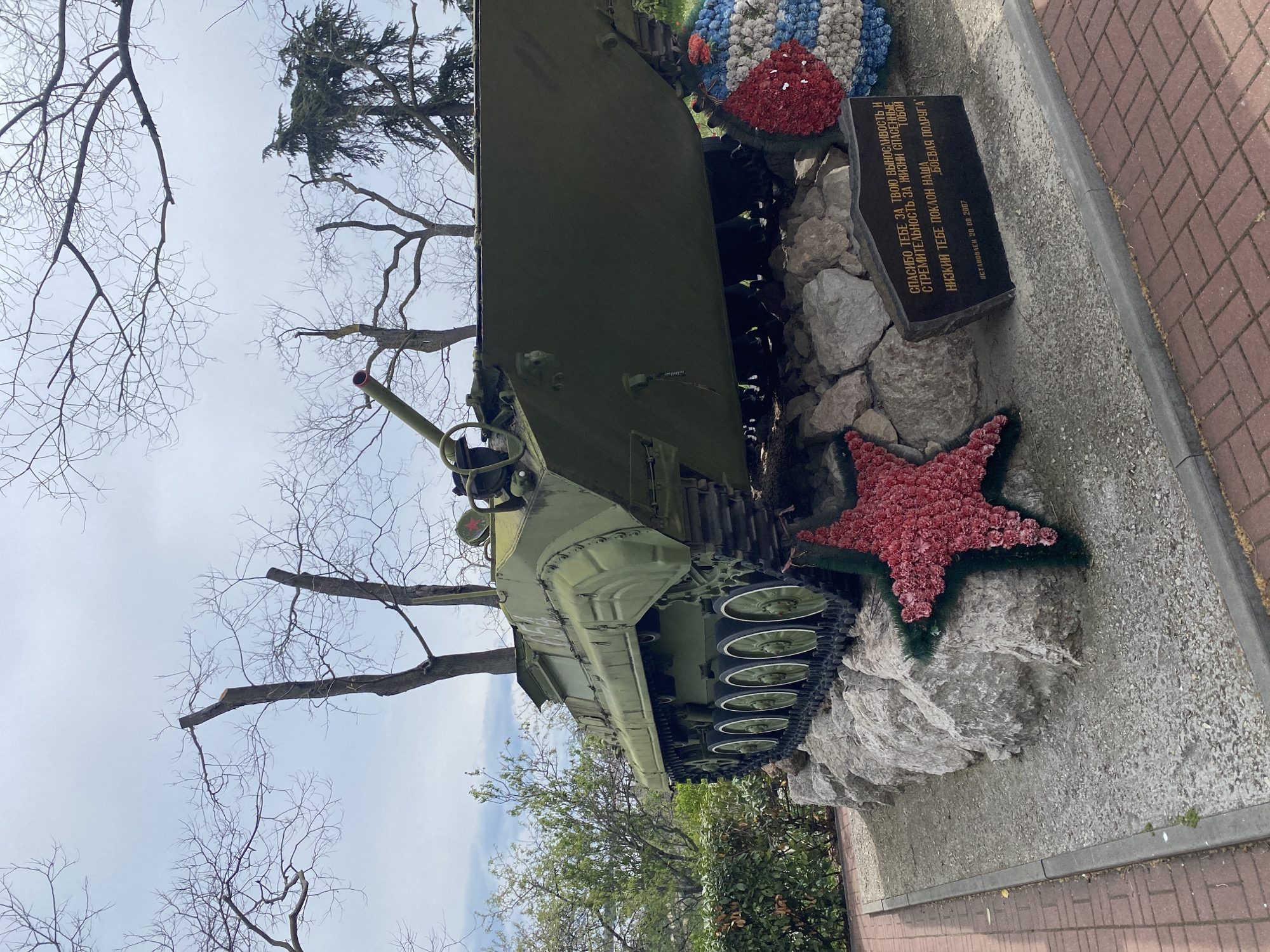 The image shows a Soviet-era armored vehicle on display in Sevastopol, Ukraine. The vehicle is painted green and mounted on a rocky base. Floral wreaths, including a red star, adorn the monument. A black plaque next to the vehicle bears an inscription in Russian, translated as: "Thank you for your endurance and speed, for the lives saved by you. Low bow to you, our fighting friend. Installed on September 20, 2007." Bare trees stand behind the vehicle, and a section of brick pavement borders the scene.