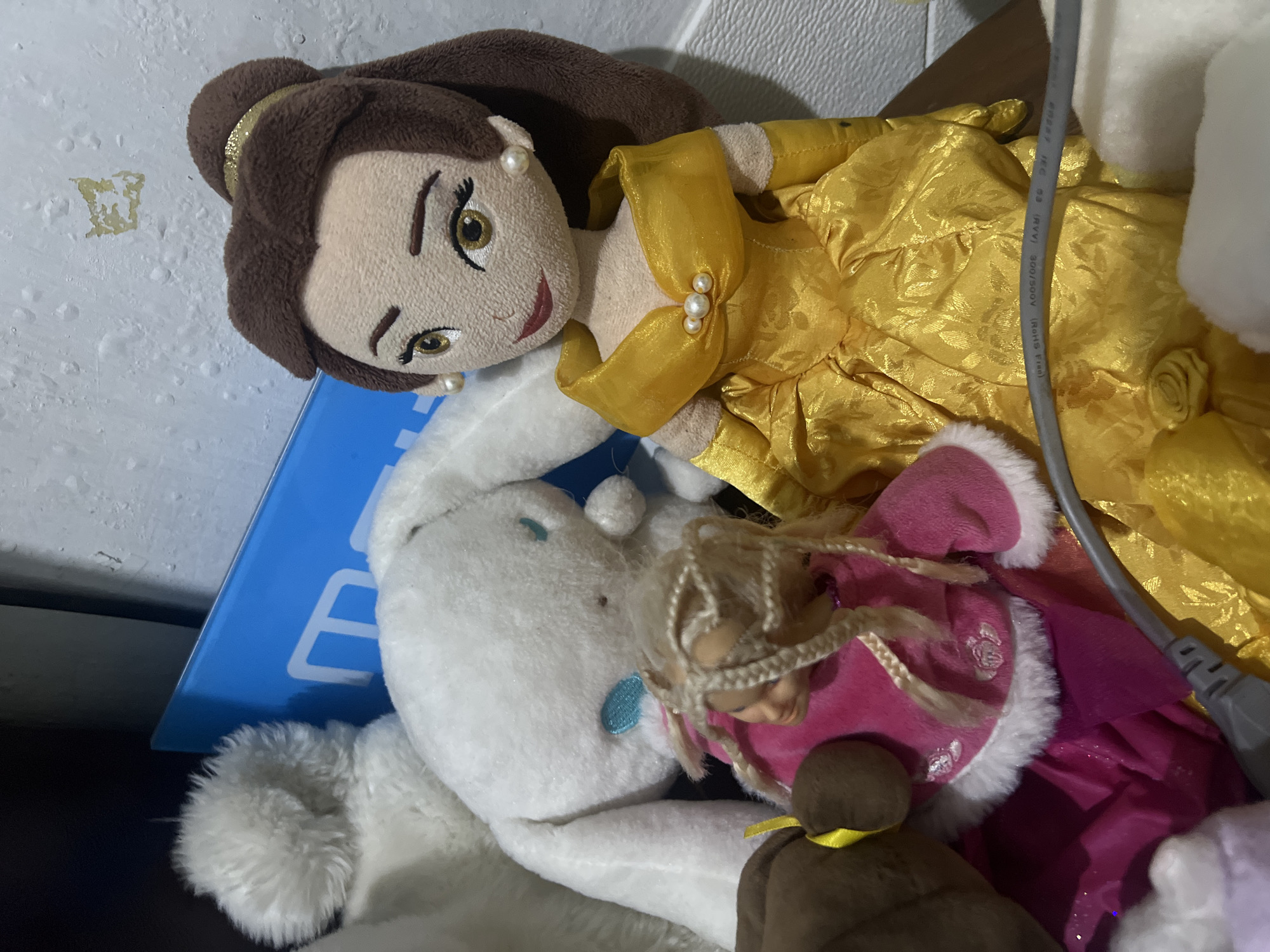In Kecamatan Pati, Indonesia, a close-up shot captures a collection of stuffed toys. A plush doll resembling Belle from Disney's "Beauty and the Beast" is positioned prominently in the foreground, adorned in her iconic yellow ballgown. To her left, a white plush bunny toy, possibly Cinnamoroll, is partially visible. In front of the bunny, a smaller doll with blonde braided hair and a pink furry jacket can be seen. The toys are arranged in front of a blue object and against a textured wall. A grey cable snakes across the lower portion of the frame, resting on the yellow fabric of Belle's dress. The lighting is dim, suggesting an indoor setting.