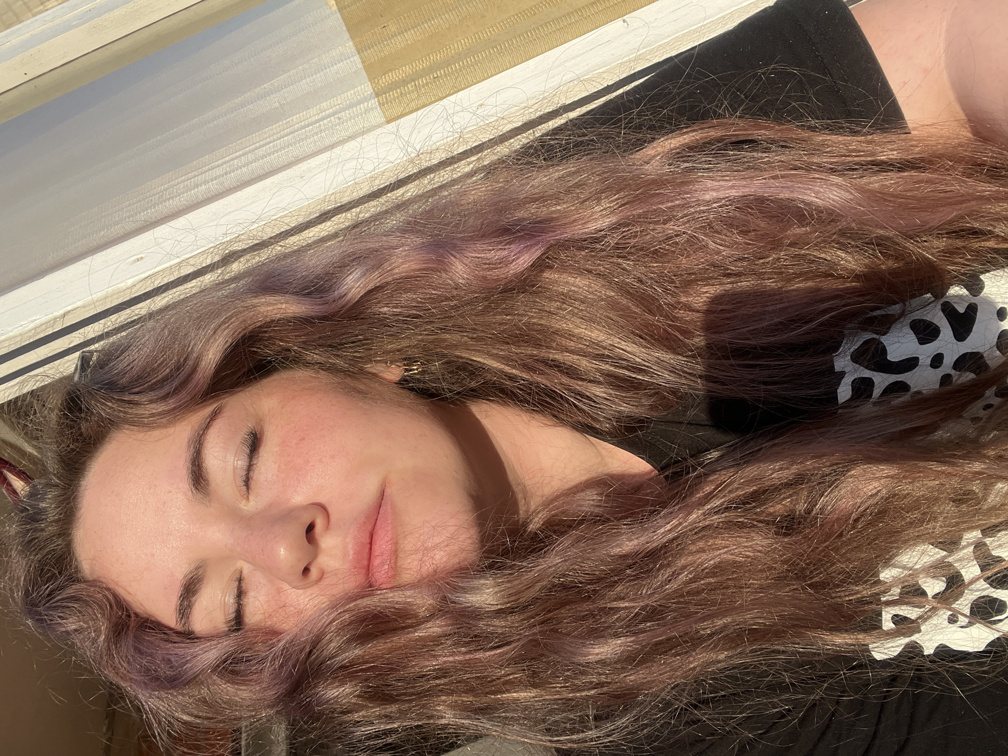 A young woman with light skin is lying down with her eyes closed. Her long, wavy brown hair with hints of purple is spread around her face. She is wearing a black top with a white and black animal print design on one shoulder. She is lying near a window with closed blinds. The lighting suggests it is daytime. There is no location information beyond what can be seen in the image. The city of Chaikovsky, Russia is not visible or discernible from the image.