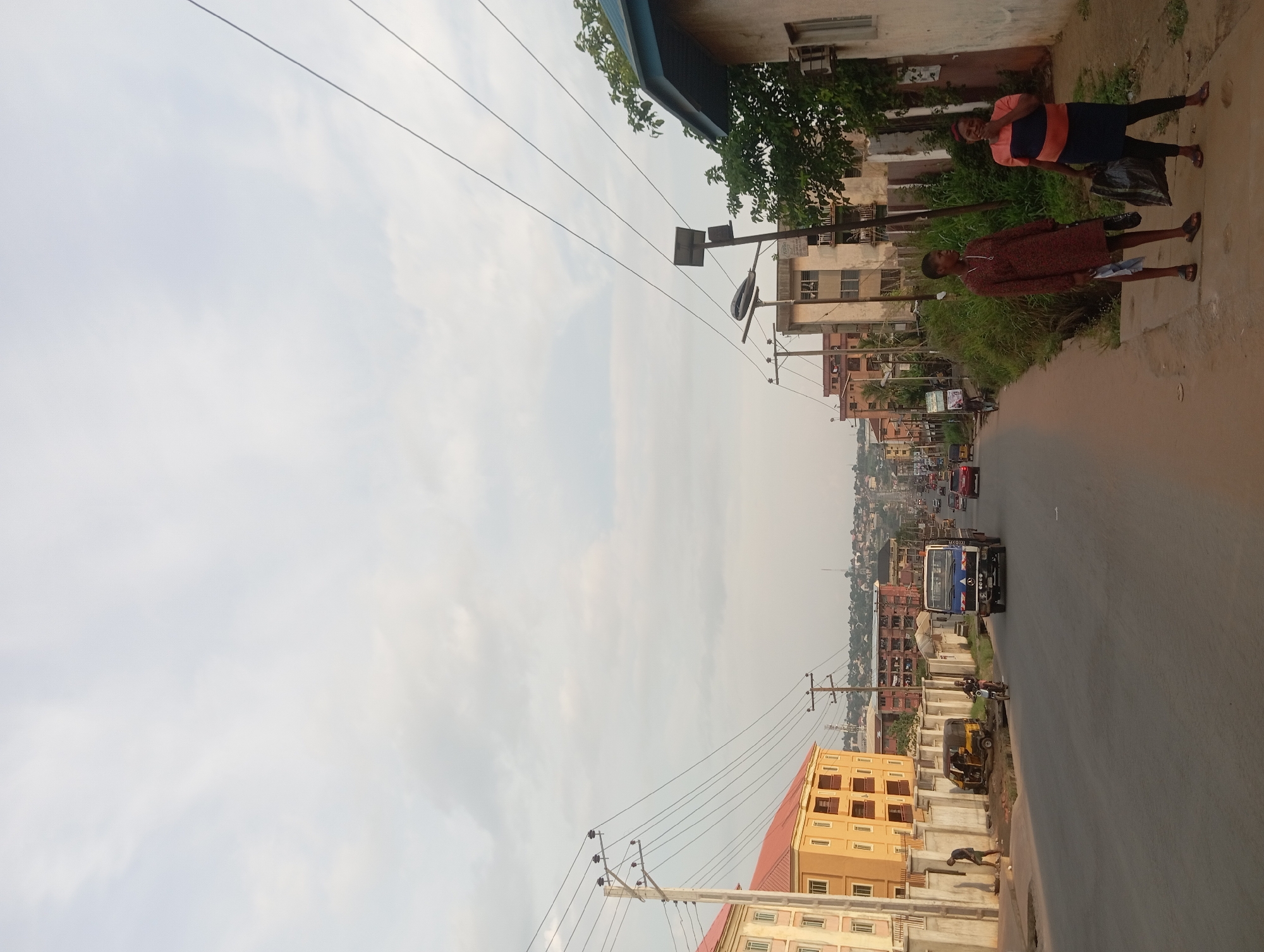 The image depicts a street scene in Nkpor, Nigeria. The view is along a paved road lined with buildings, utility poles, and power lines. The sky is overcast. Two women are walking along the side of the road in the foreground. A blue and white bus is on the road, along with other vehicles further down the street. A building with a red roof is located on the left side of the road in the foreground. A tricycle is parked by the side of the road. A man is crouched on the side of the road.