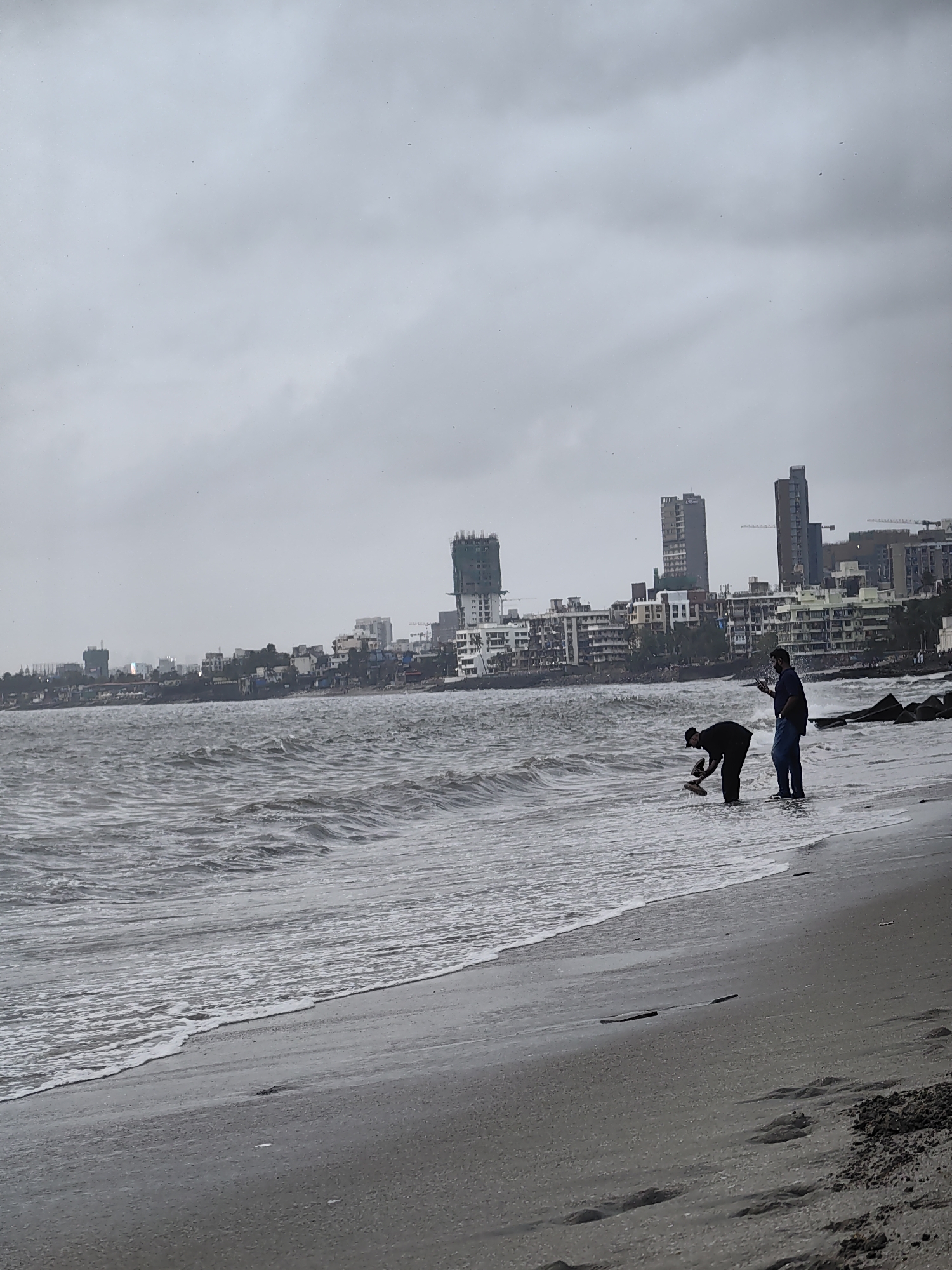 The image depicts a cloudy day at a beach in Mumbai, India, with the city skyline visible in the background. The sea is rough, with waves crashing onto the shore. In the foreground, two individuals are present on the beach. One person, wearing dark clothing and a cap, is bent over near the water, possibly picking something up from the sand or sea. Another person, standing upright and wearing a dark shirt and blue jeans, appears to be looking at their mobile phone. The beach itself is sandy, with some debris and footprints visible. The distant cityscape features numerous buildings, including skyscrapers and lower-rise structures, suggesting a densely populated urban area. The overall mood of the image is somber due to the overcast sky and the choppy sea.