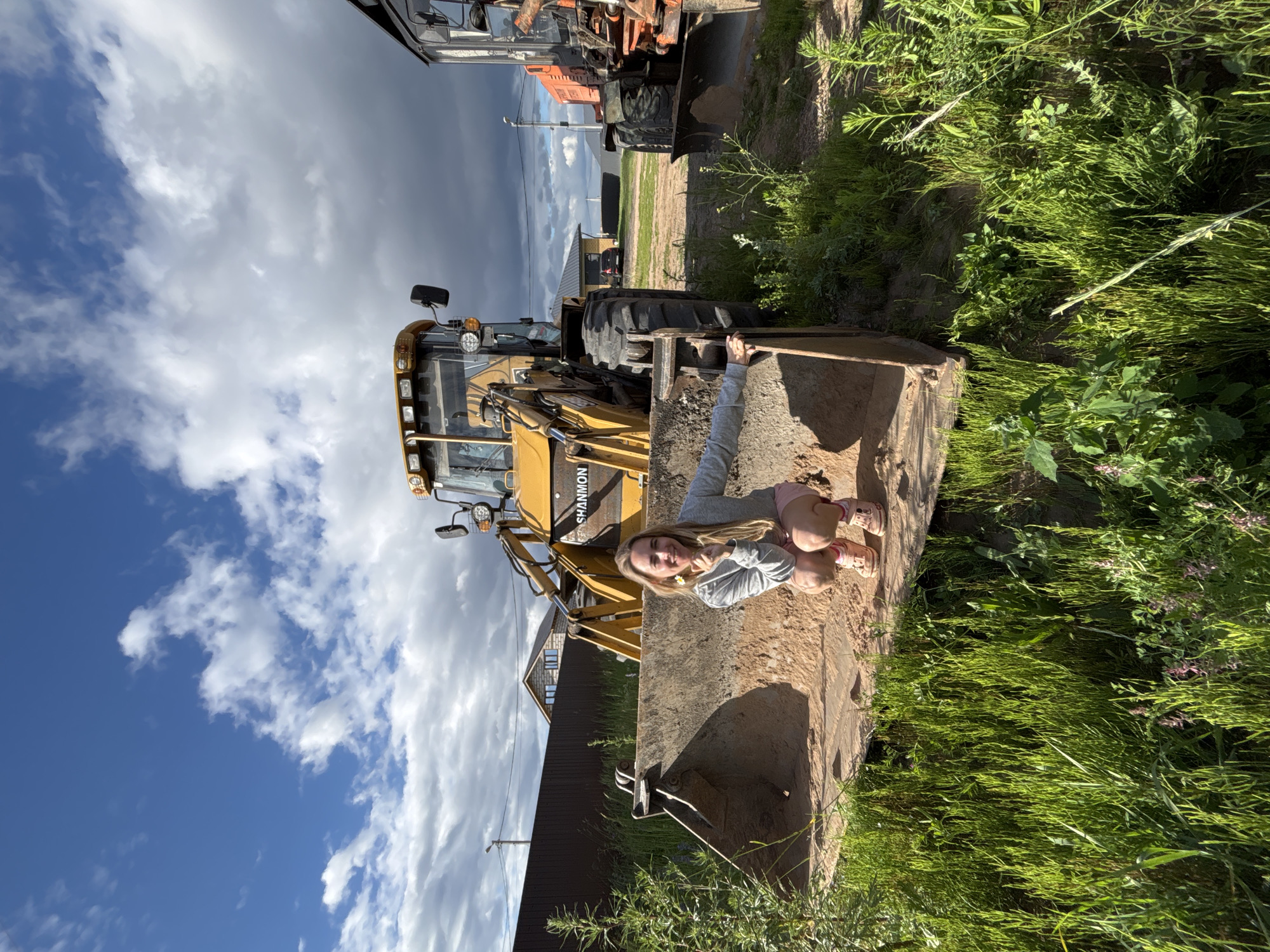 A young girl sits on the edge of a large, dirt-filled bucket of a yellow excavator. She is wearing a gray sweatshirt and pink shorts, and her feet are clad in pink sandals. The girl is smiling and holding a small white flower. Behind her, the excavator is parked on a patch of ground surrounded by lush green grass and vegetation. Further in the background, a dark brown wooden fence runs horizontally, with a house and a pole visible behind it. The sky above is a mixture of bright blue and dramatic, dark clouds, suggesting a dynamic weather system. To the left of the yellow excavator, another large piece of orange construction equipment is visible, indicating a work or construction site. The overall scene appears to be outdoors during the daytime. The text "SHANNON" is visible on the side of the yellow excavator. The location context is Chaikovsky, Russia.