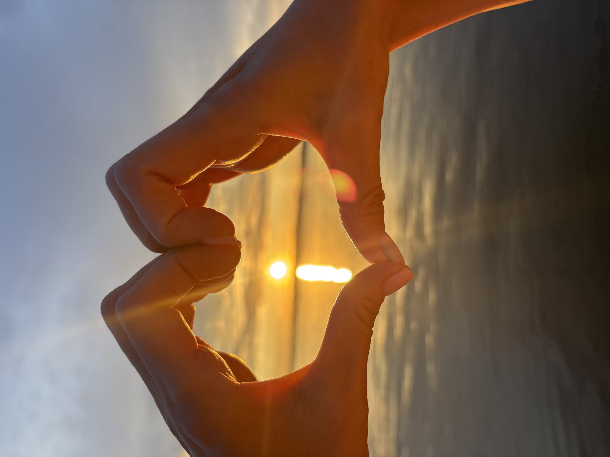 A close-up, vertical shot captures two hands forming a heart shape against a sunset over water. The sun, a bright orb, is positioned centrally within the heart, casting a golden reflection on the calm surface of a body of water. The sky transitions from a pale blue at the top to warm oranges and yellows near the horizon, with hints of clouds. The hands, illuminated by the sunset, are positioned in the foreground, with the left hand's thumb and forefinger touching the right hand's forefinger and thumb. The water appears dark and reflective, with subtle ripples. The scene is devoid of any specific landmarks that would identify it as being in Chaikovsky, Russia.
