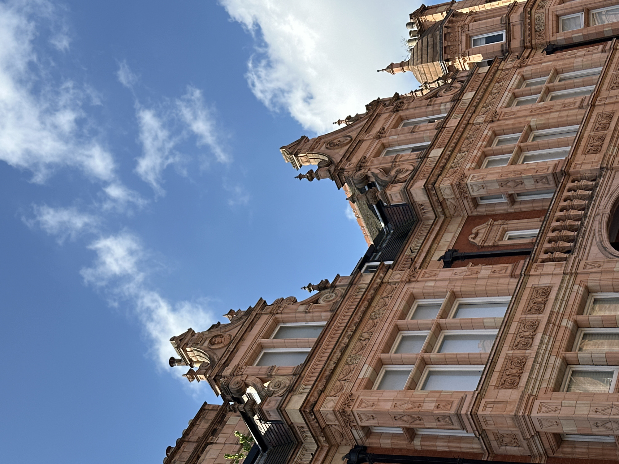 The image shows an upward-tilted view of a richly ornamented building in London, United Kingdom, against a blue sky with scattered white clouds. The building has a light reddish-brown facade with ornate carvings and multiple windows arranged in rows. Gargoyles and decorative stonework adorn the upper portions of the building. A glimpse of what appears to be a rooftop or upper-level structure is visible at the top of the frame.