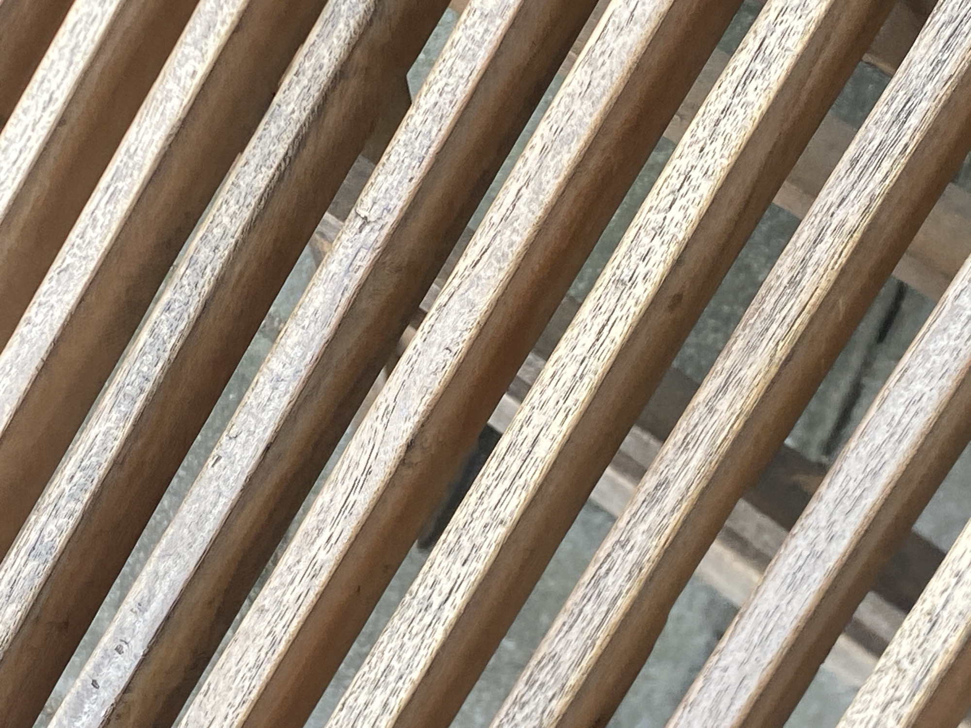 The image shows a close-up, angled view of weathered wooden slats, likely part of a bench or outdoor furniture. The wood has a natural brown tone with visible grain and some lighter, bleached areas suggesting exposure to the elements. The slats are arranged parallel to each other with narrow gaps between them. In the background, blurred glimpses of a greyish, textured surface, possibly pavement or another outdoor material, can be seen. The scene appears to be outdoors, and while the exact location in Mengwi, Indonesia, cannot be definitively determined from the image alone, the context suggests an urban or semi-urban outdoor setting. There are no people or discernible actions visible in the image.