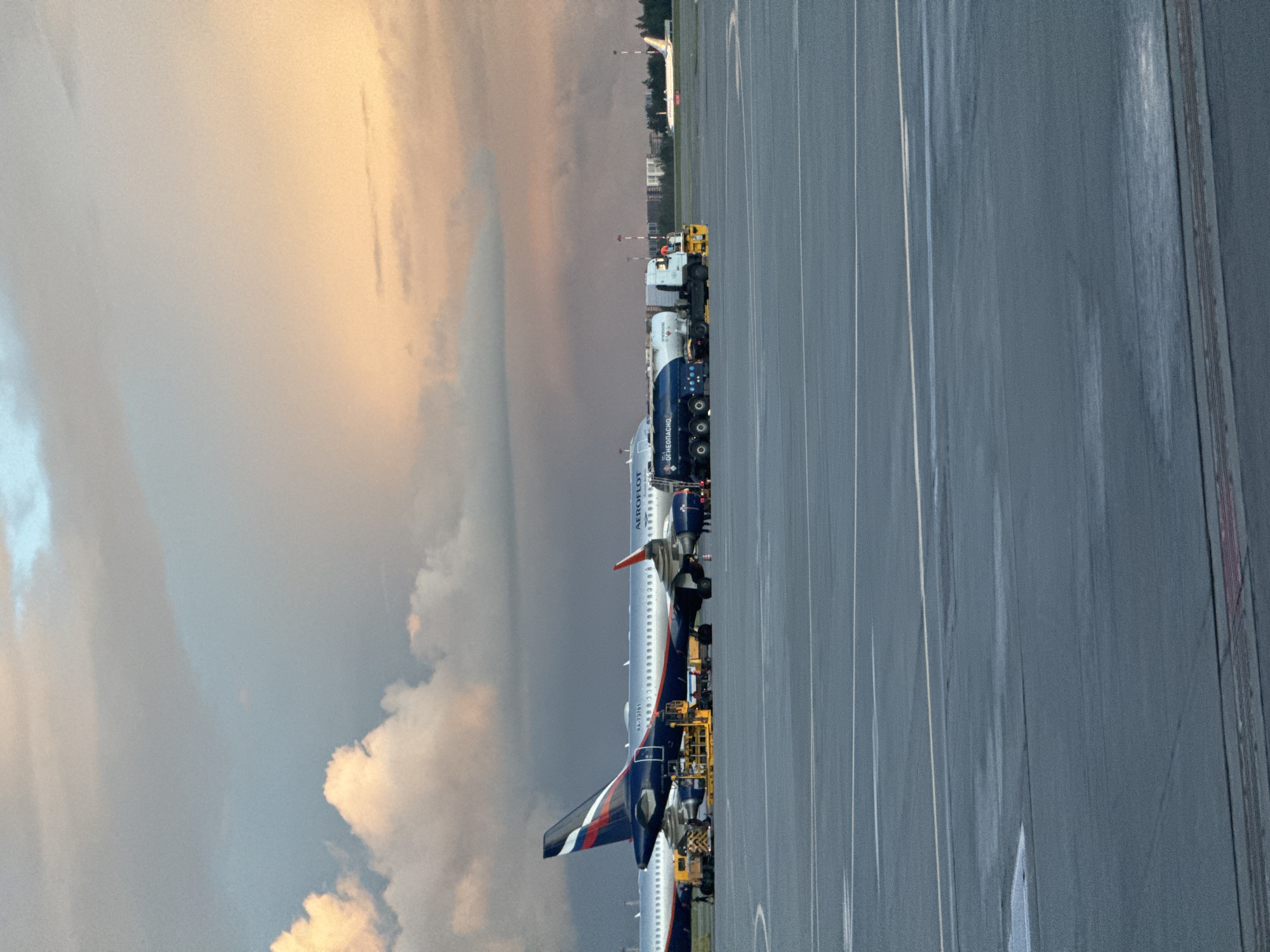 The image shows an Aeroflot passenger jet parked on an airport tarmac, likely at Sheremetyevo International Airport, given its location in Khimki, Russia. The plane is being serviced, as evidenced by the presence of yellow maintenance vehicles. A line of refueling trucks is visible behind the plane, indicating that it is likely being prepared for its next flight. The sky is overcast with hues of pink and grey. The tarmac appears wet, possibly from recent rain. There are buildings and trees visible in the background, suggesting a suburban or industrial area. There are no people immediately visible in the frame, although their presence is implied by the maintenance vehicles.