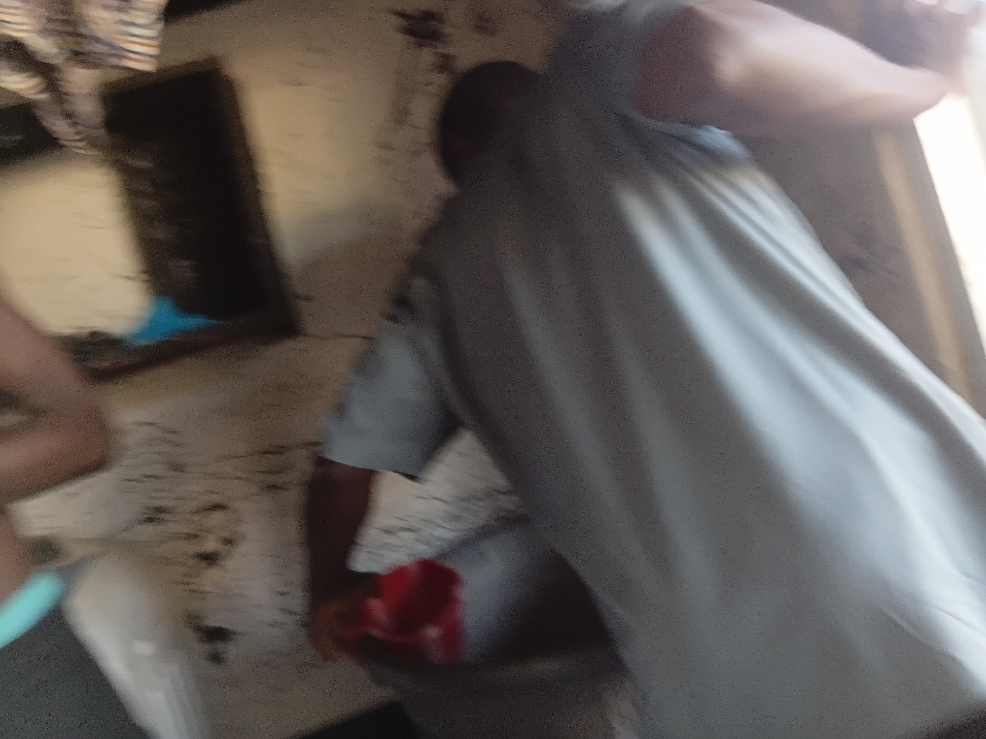 The image, taken in Jalingo, Nigeria, depicts a dimly lit interior space with textured walls, possibly made of cement or plaster, showing some dirt and debris. A person, whose face is obscured by motion blur and their body is partially visible, is wearing a light-colored garment. They are leaning over a large, dark basin or container. In their hand, they are holding a small red object, possibly a cup or scoop. To the left of the frame, another person's arm and hand are visible, with a light blue object also present. The overall impression is one of a functional, perhaps utilitarian, environment where a task is being performed. The blur suggests movement and possibly a fast shutter speed.