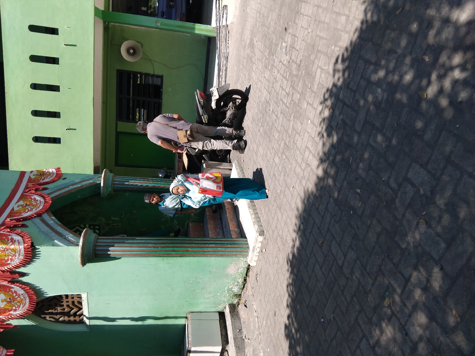 In the city of Kepuh Kiriman, Indonesia, on a sunny day, a group of people are gathered outside a green building. A young girl, wearing a light blue hijab and a teal abaya, stands near the entrance, holding a red and white bag. Beside her, a boy in a light blue uniform and a dark cap stands on the steps. To the right, a man wearing a grey t-shirt and pants, with a brown bag slung across his shoulder, stands next to a black motorcycle. The building, painted a vibrant green, features decorative red and white banners above the entrance. The ground is paved with interlocking grey bricks, and shadows from trees dapple the scene.